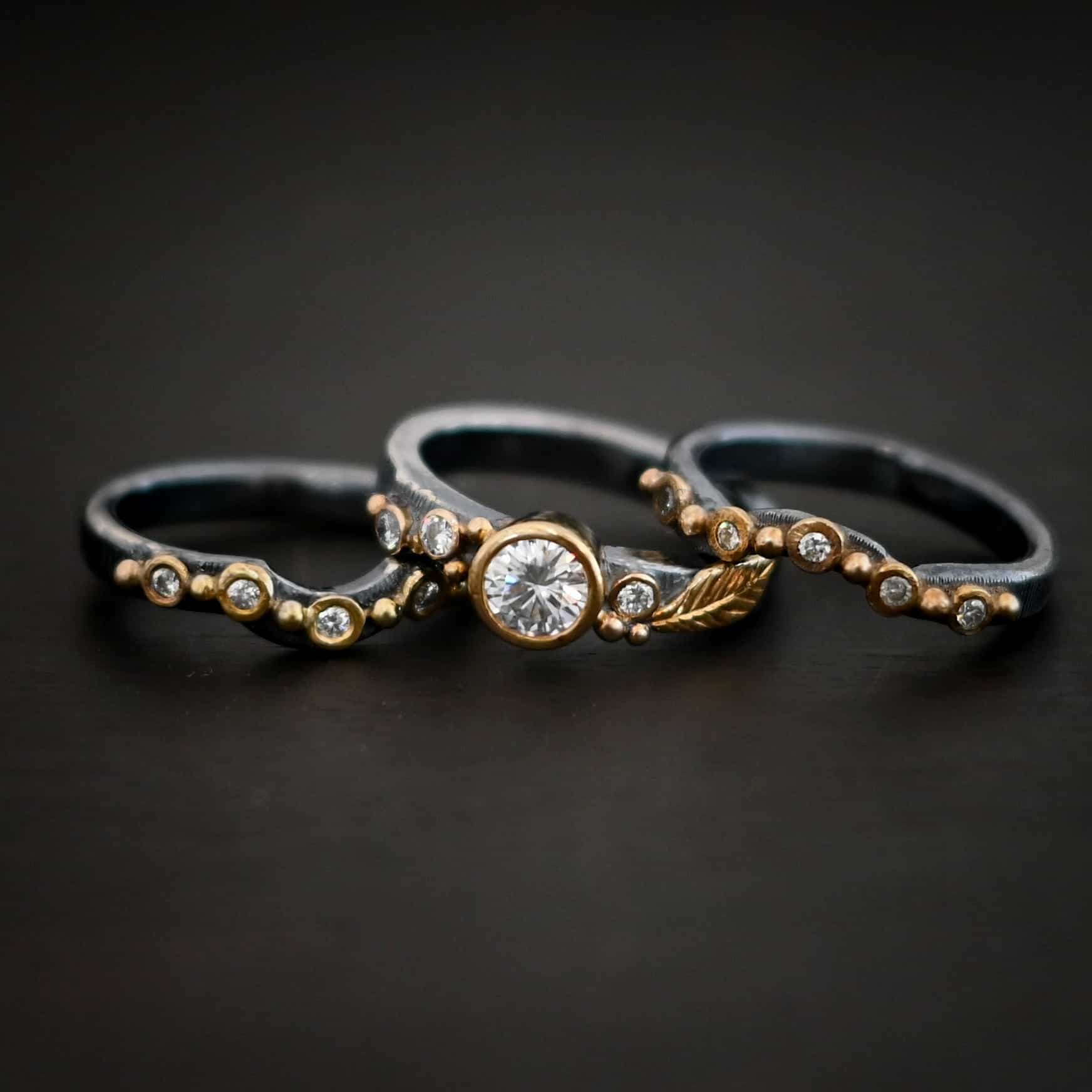 Photo of the "Forever Held" Ring. Ring has a 1.5 carat moissanite in the center, with two diamonds on the left side of it and one on the right. There is gold ball detail and a gold leaf on the band as well. Ring has a top and bottom band that have a dip in the center to fit around the center stone. These bands have multiple diamonds around them with balls of gold that separate each diamond. Ring is sitting against a dark background.