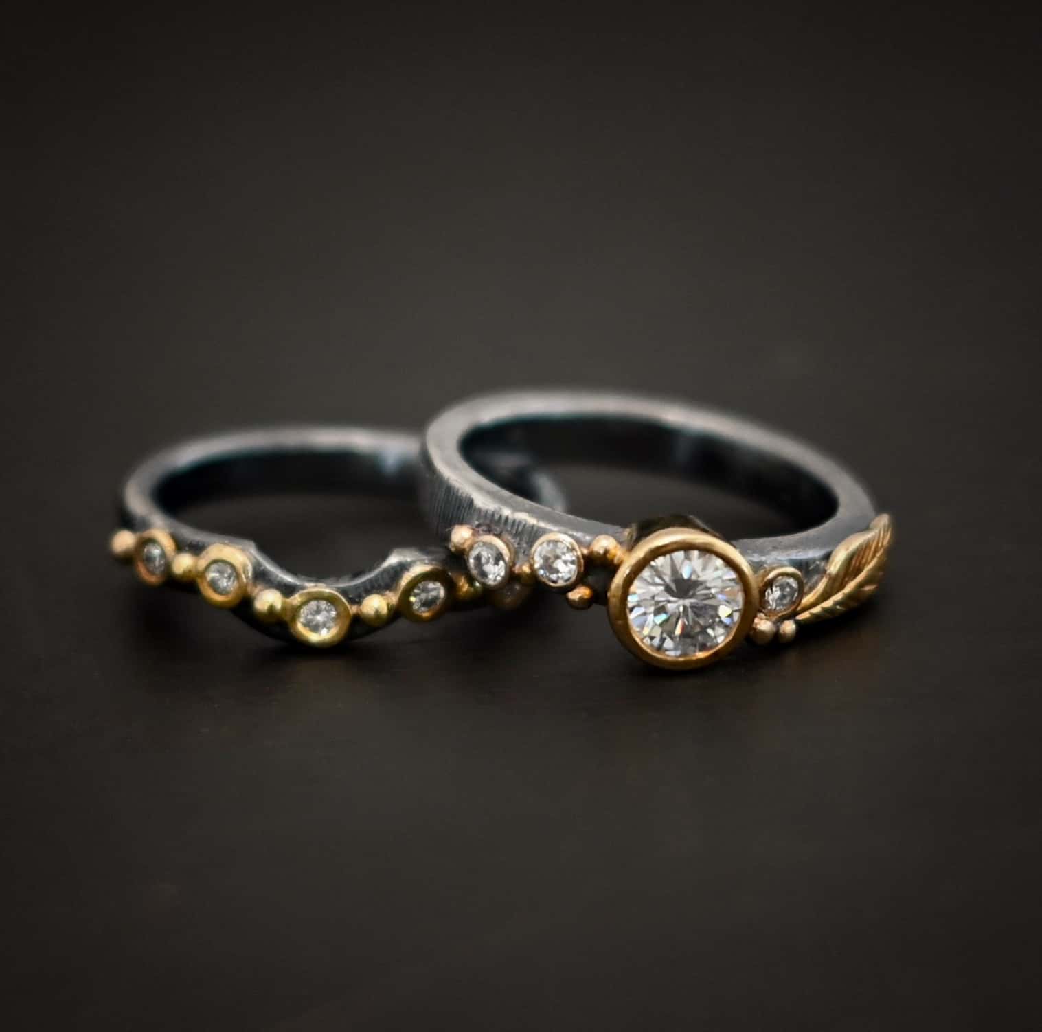 Photo of the "Forever Held" Ring. Ring has a 1.5 carat moissanite in the center, with two diamonds on the left side of it and one on the right. There is gold ball detail and a gold leaf on the band as well. Ring has a bottom band that has a dip in the center to fit around the center stone. This band has multiple diamonds around it with balls of gold that separate each diamond. Ring is sitting against a dark background.