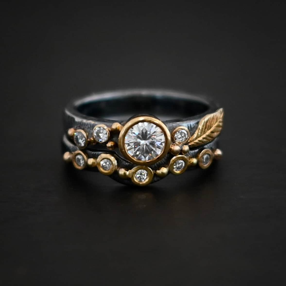 Photo of the "Forever Held" Ring. Ring has a 1.5 carat moissanite in the center, with two diamonds on the left side of it and one on the right. There is gold ball detail and a gold leaf on the band as well. Ring has a bottom band that has a dip in the center to fit around the center stone. This band has multiple diamonds around it with balls of gold that separate each diamond. Ring is sitting against a dark background.