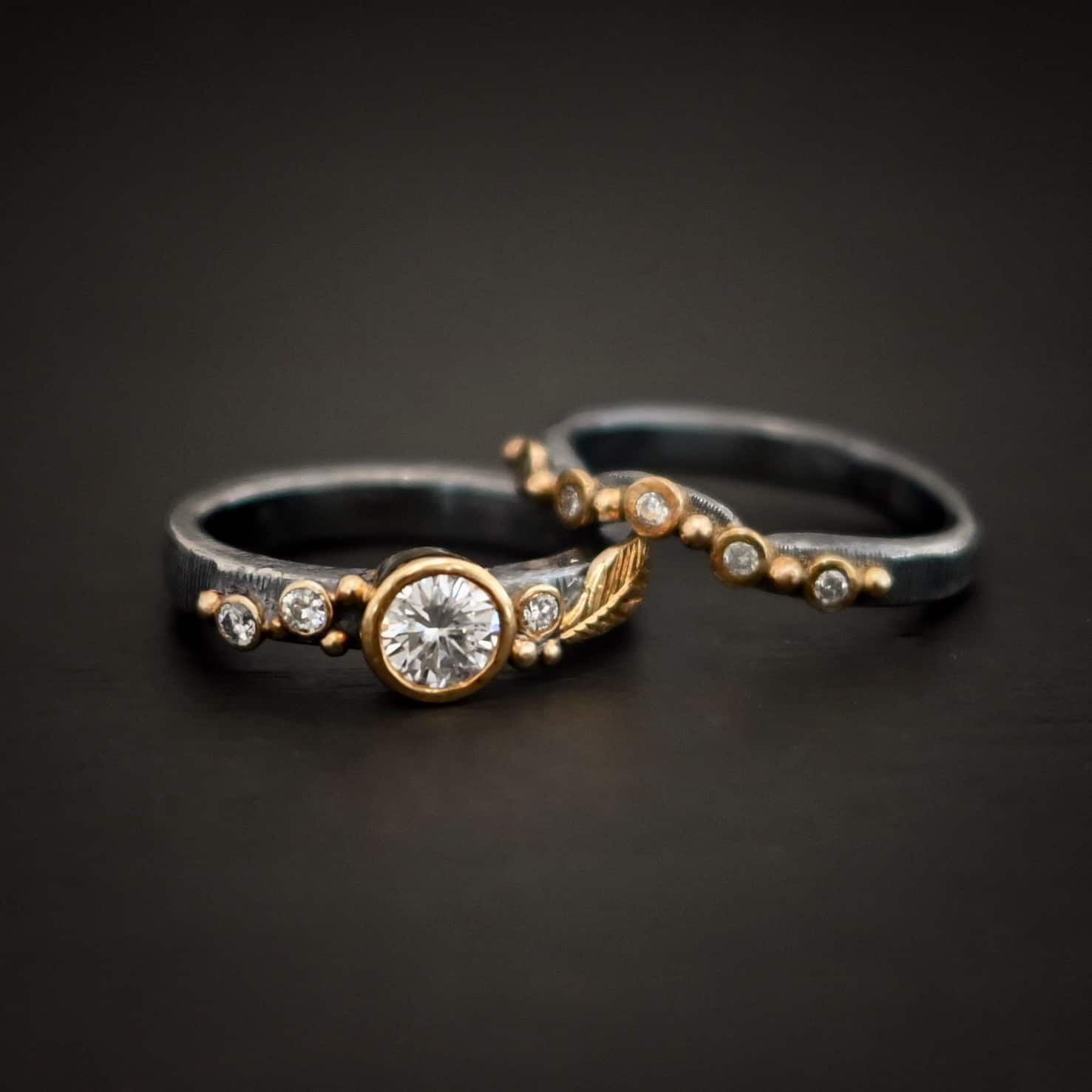 Photo of the "Forever Held" Ring. Ring has a 1.5 carat moissanite in the center, with two diamonds on the left side of it and one on the right. There is gold ball detail and a gold leaf on the band as well. Ring has a top band that has a dip in the center to fit around the center stone. This band has multiple diamonds around it with balls of gold that separate each diamond. Ring is sitting against a dark background.