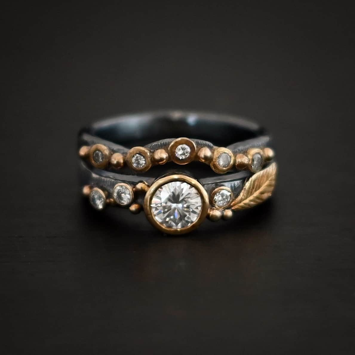Photo of the "Forever Held" Ring. Ring has a 1.5 carat moissanite in the center, with two diamonds on the left side of it and one on the right. There is gold ball detail and a gold leaf on the band as well. Ring has a top band that has a dip in the center to fit around the center stone. This band has multiple diamonds around it with balls of gold that separate each diamond. Ring is sitting against a dark background.