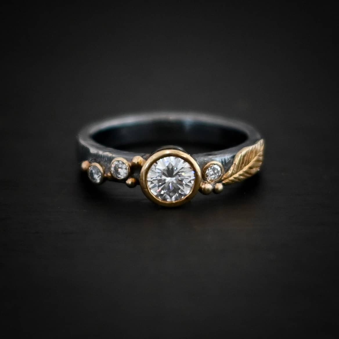 Photo of the "Forever Held" Ring. Ring has a 1.5 carat moissanite in the center, with two diamonds on the left side of it and one on the right. There is gold ball detail and a gold leaf on the band as well. Ring is sitting against a dark background.