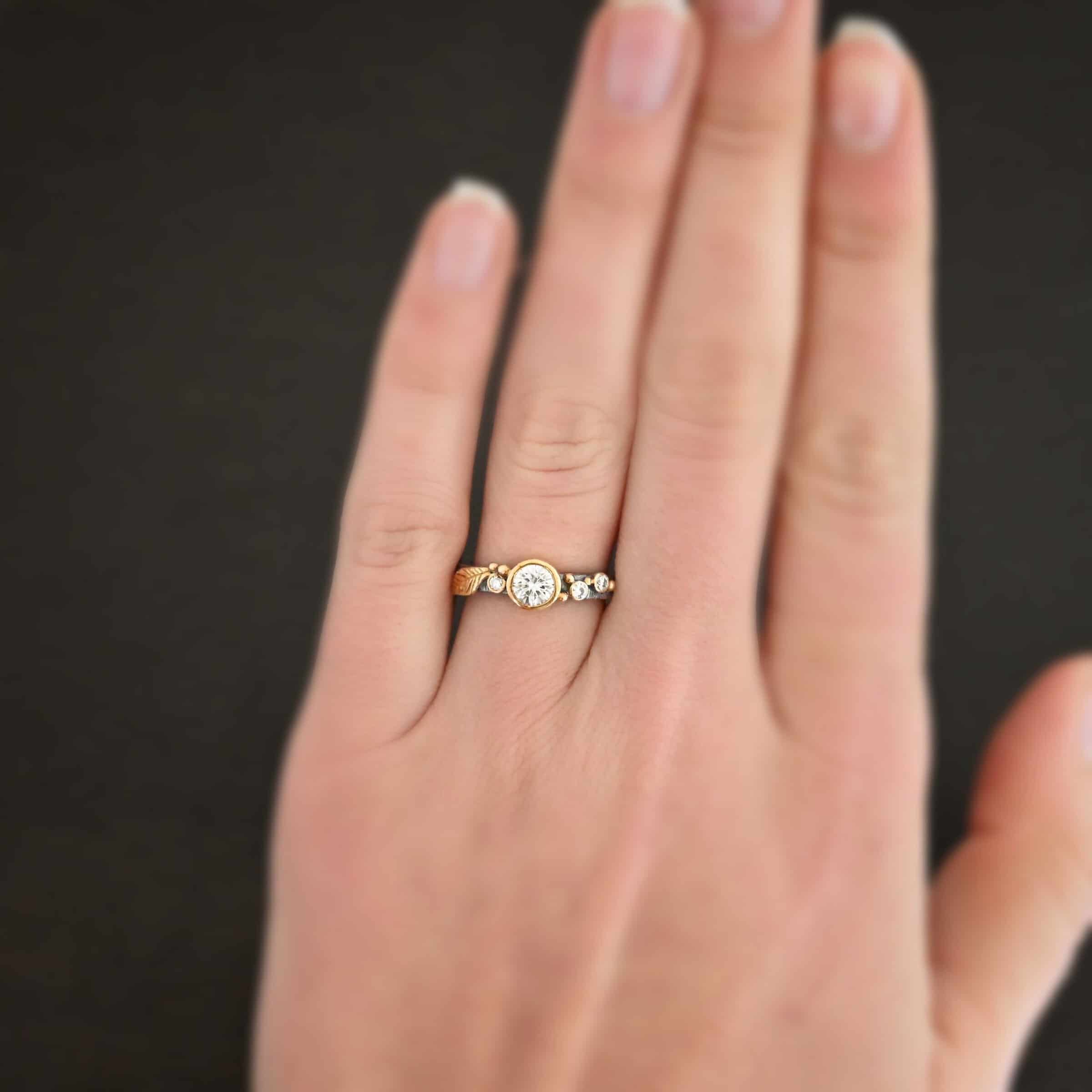 Photo of the "Forever Held" Ring. Ring has a 1.5 carat moissanite in the center, with two diamonds on the right side of it and one on the left. There is gold ball detail and a gold leaf on the band as well. Ring is worn by a model.