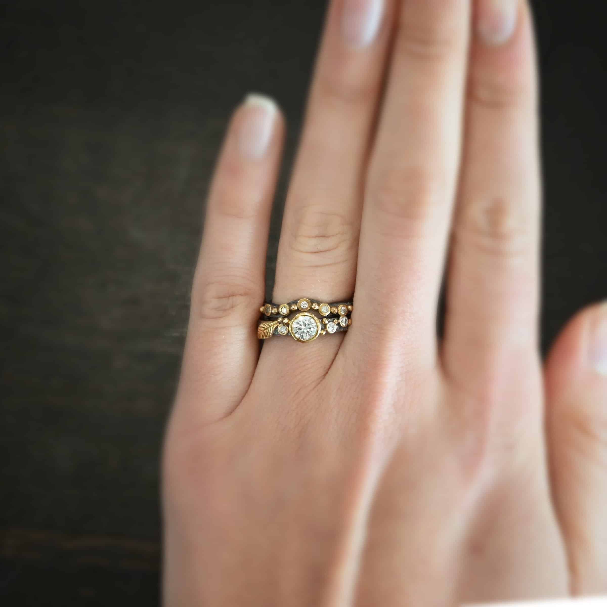 Photo of the "Forever Held" Ring. Ring has a 1.5 carat moissanite in the center, with two diamonds on the right side of it and one on the left. There is gold ball detail and a gold leaf on the band as well. The top band has a dip in the middle to go around the center stone and fit with it. The band has multiple diamonds and gold ball details on it. Ring is worn by a model.