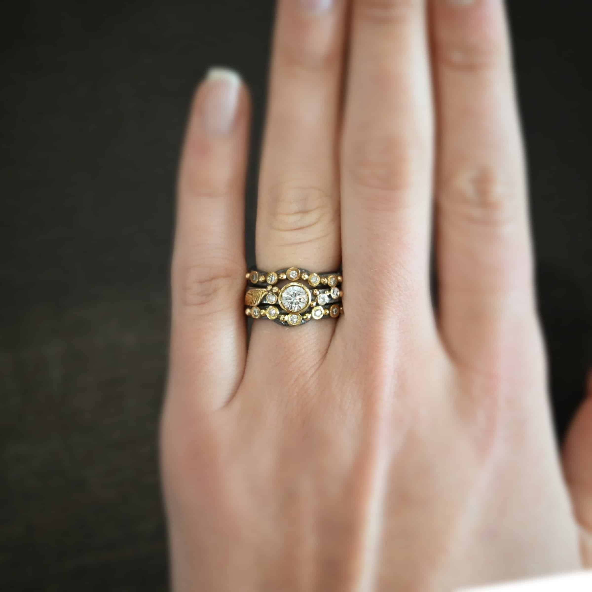Photo of the "Forever Held" Ring. Ring has a 1.5 carat moissanite in the center, with two diamonds on the right side of it and one on the left. There is gold ball detail and a gold leaf on the band as well. The top and bottom band both have a dip in the middle to go around the center stone and fit with it. These bands both have multiple diamonds and gold ball detail on them. Ring is worn by a model.
