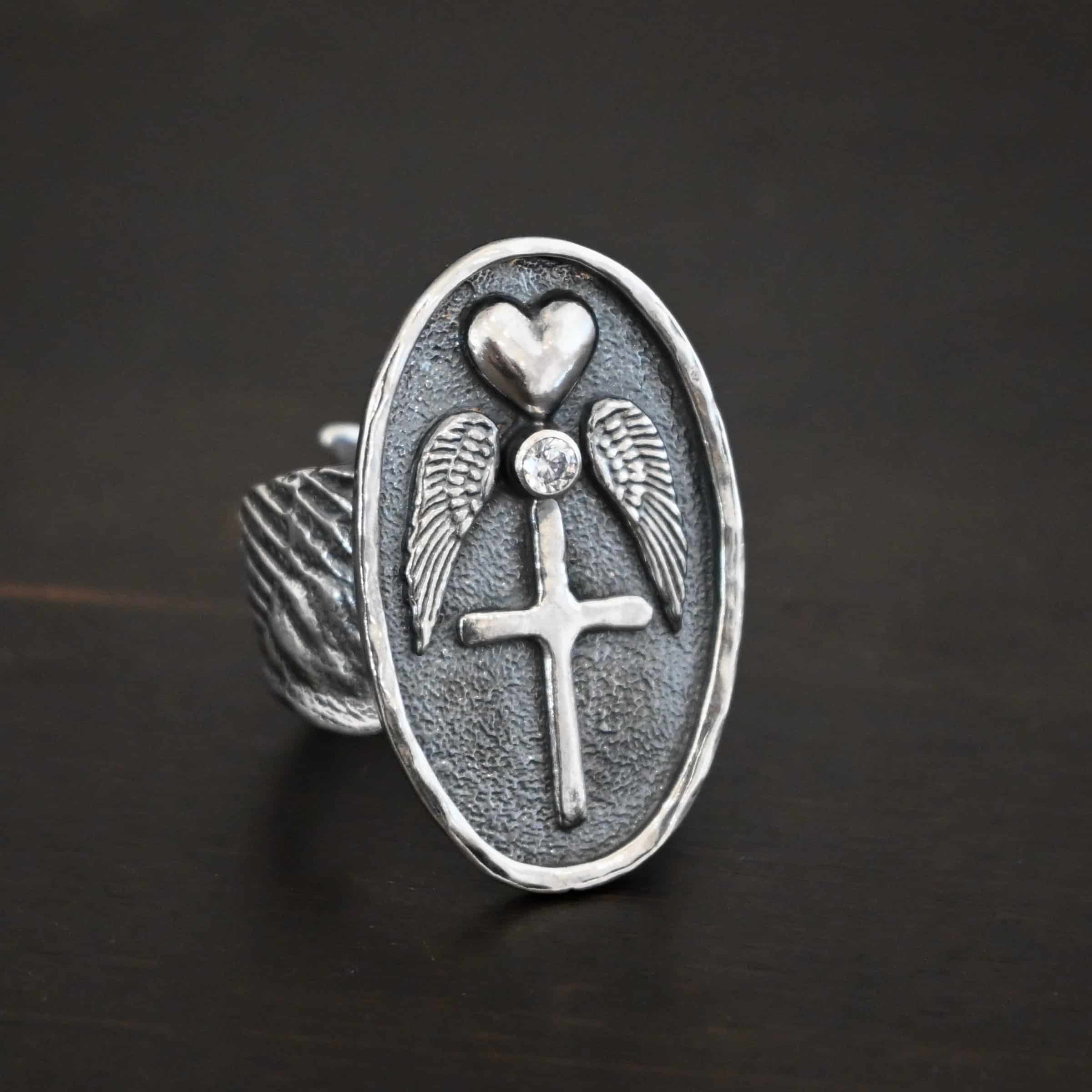 My Forever Angel Ring CZ Photo of "My Forever Angel" Cremation Ash Ring. Ring is made out of sterling silver. It is a large oval with textured sterling silver on the inside and a line of sterling silver that goes around the edges of the oval that is shiny. The inside of the ring has a heart charm on the top, a cz beneath it on the tip, two angel wings beneath the heart, one on each side of it, and a cross beneath the cz, with the tips of each angel wing touching the left side of the cross and the right side. The ring band is made out of thick sterling silver angel wings. One angel wing is beneath the other, so they overlap, but don't touch. Ring is sitting against a dark background.