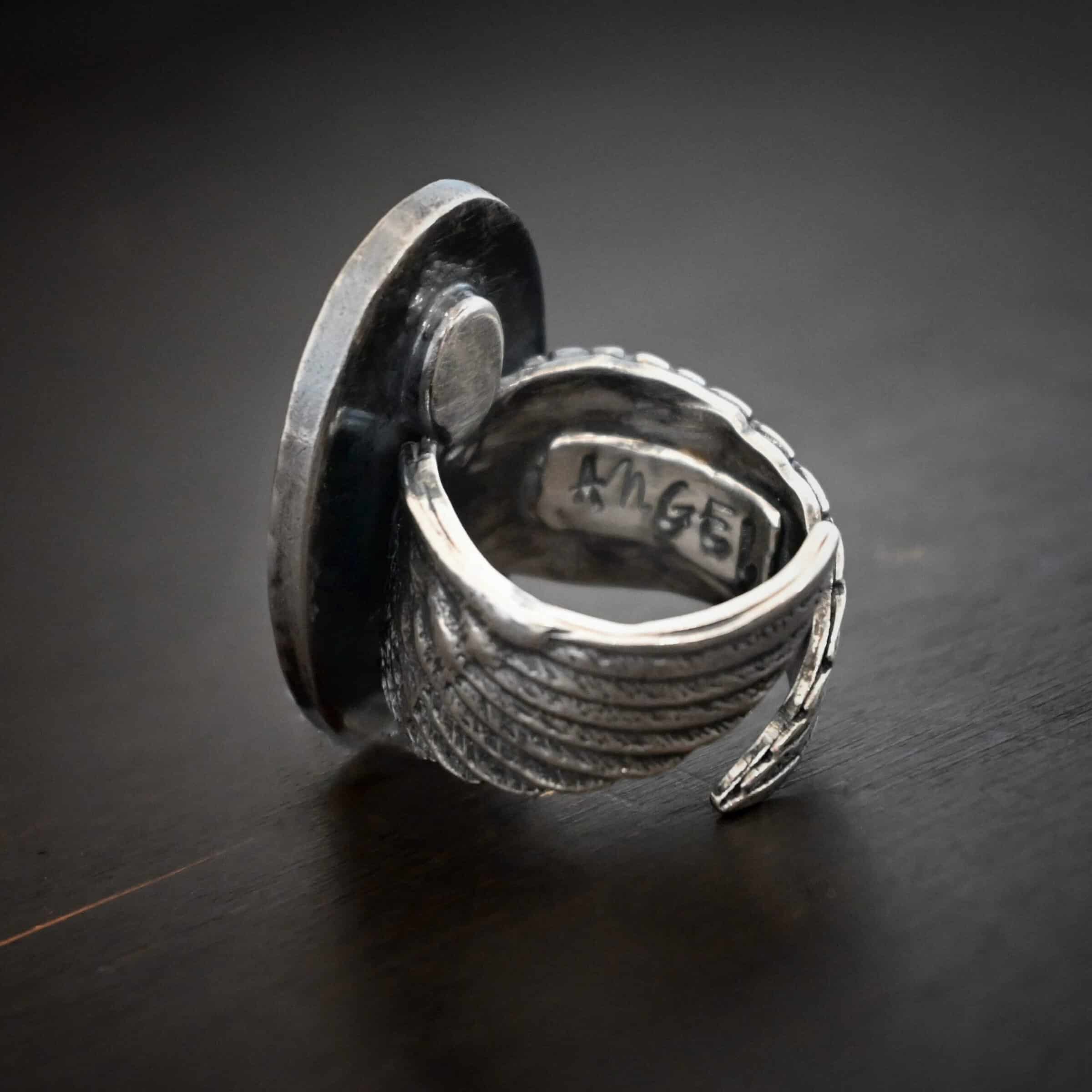 My Forever Angel Cremation ring Photo of the back of the "My Forever Angel" Cremation Ash Ring. Ring is made out of sterling silver. It is a large oval with textured sterling silver on the inside and a line of sterling silver that goes around the edges of the oval that is shiny. The ring band is made out of thick sterling silver angel wings. One angel wing is beneath the other, so they overlap, but don't touch. There is a small shiny square on the inside of the ring with the word "Angel" stamped into it. Ring is sitting against a dark background.