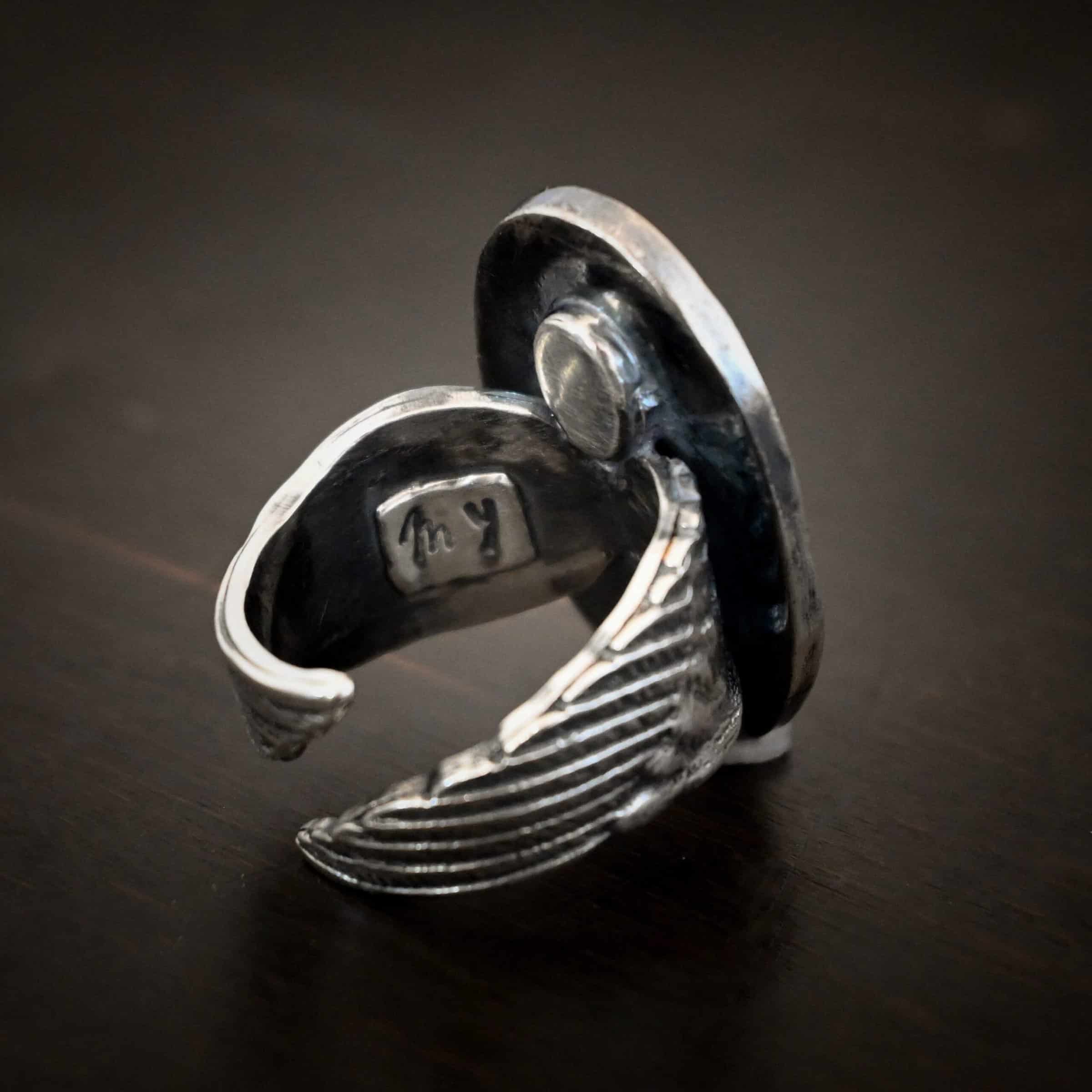 My Forever Angel Cremation ring Photo of the back of the "My Forever Angel" Cremation Ash Ring. Ring is made out of sterling silver. It is a large oval with textured sterling silver on the inside and a line of sterling silver that goes around the edges of the oval that is shiny. The ring band is made out of thick sterling silver angel wings. One angel wing is beneath the other, so they overlap, but don't touch. There is a small shiny square on the inside of the ring with the word "My" stamped into it. Ring is sitting against a dark background.