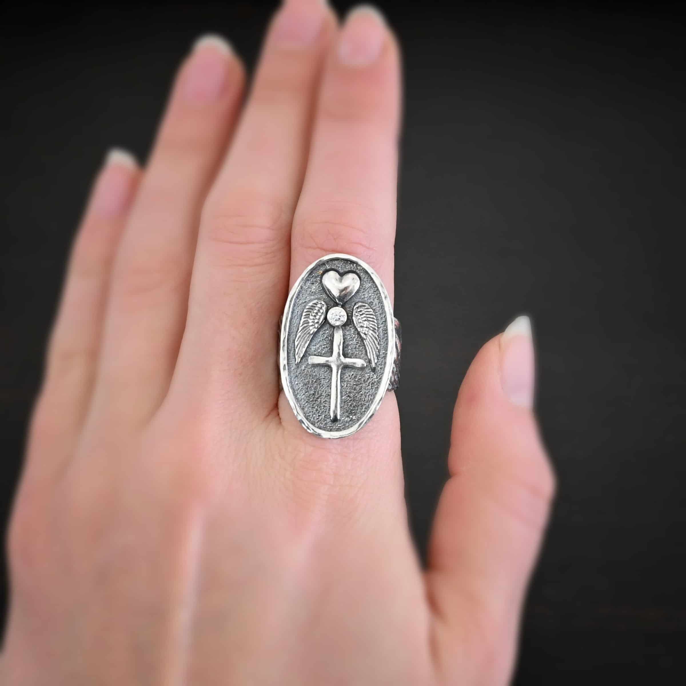 My Forever Angel Ring CZ Photo of "My Forever Angel" Cremation Ash Ring. Ring is made out of sterling silver. It is a large oval with textured sterling silver on the inside and a line of sterling silver that goes around the edges of the oval that is shiny. The inside of the ring has a heart charm on the top, a diamond beneath it on the tip, two angel wings beneath the heart, one on each side of it, and a cross beneath the diamond, with the tips of each angel wing touching the left side of the cross and the right side. The ring band is made out of thick sterling silver angel wings. One angel wing is beneath the other, so they overlap, but don't touch. Ring is worn by a model.