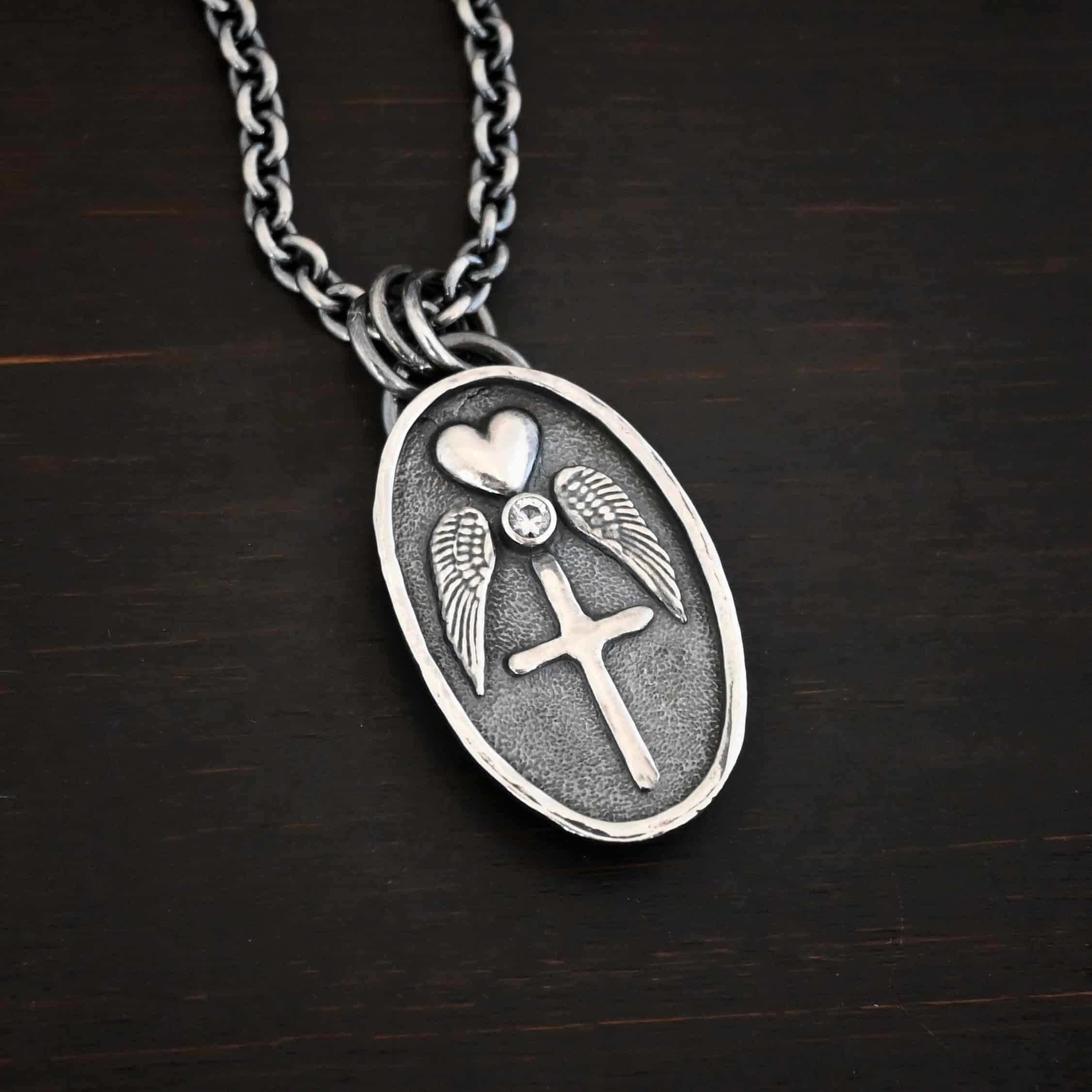 Photo of the "My Forever Angel Cremation Ash" Necklace. Made out of sterling silver. The necklace has a large, oval shaped, thick piece of silver for the pendant. On the inside of the oval is a small heart at the top that sticks up so it is noticeable. There is a small diamond on the bottom tip of the heart. There is an angel wing beneath the heart on each side, surrounding the diamond. Each wing faces outwards. Beneath the diamond is a cross that is soldered. The end of each angel wing touches the left and right side of the cross. There is a thick silver loop soldered to the top. The chain of the necklace goes through the loop. Necklace is lying against a dark background.