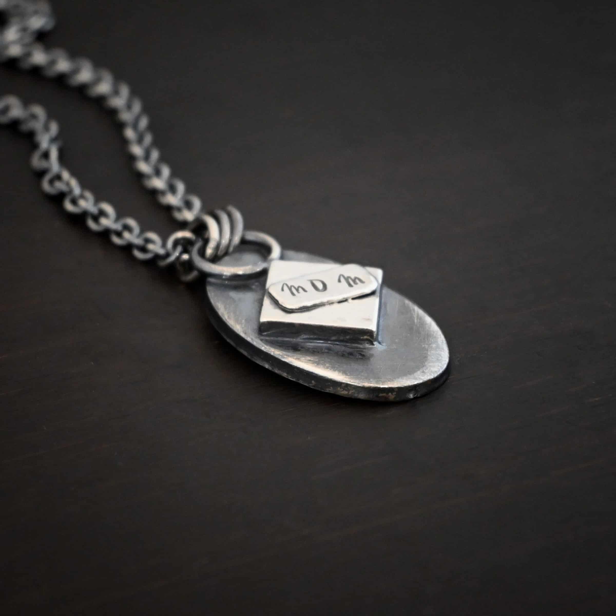 Photo of the back of the "My Forever Angel Cremation Ash" Necklace. Made out of sterling silver. The necklace has a large, oval shaped, thick piece of silver for the pendant. There is a large, shiny piece of diamond shaped silver that takes up most of the back. In the center, there is a smaller, rectangle piece of silver with the word "Mom" stamped into it. Necklace is lying against a dark background. Necklace is lying against a dark background.