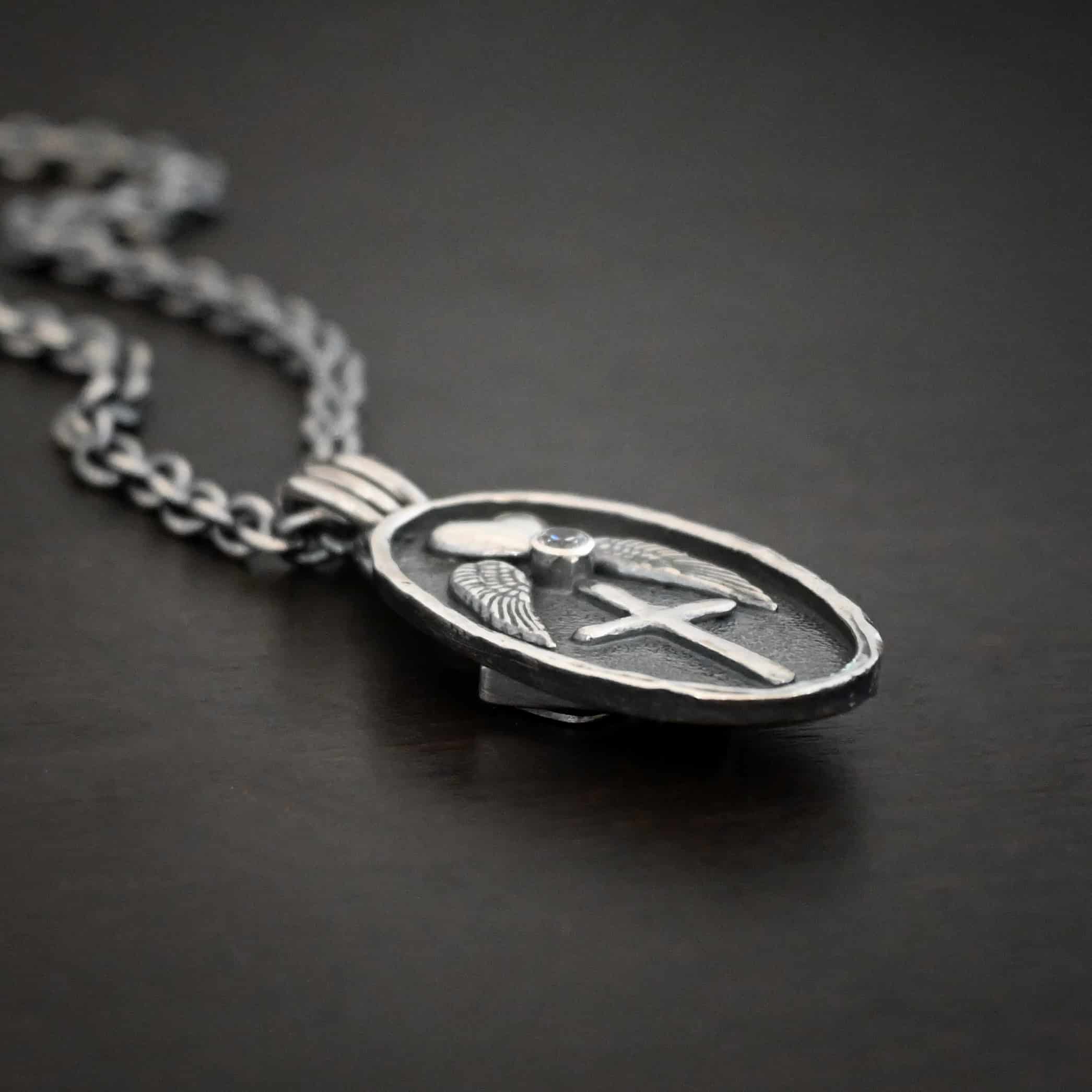Photo of the "My Forever Angel Cremation Ash" Necklace. Made out of sterling silver. The necklace has a large, oval shaped, thick piece of silver for the pendant. On the inside of the oval is a small heart at the top that sticks up so it is noticeable. There is a small diamond on the bottom tip of the heart. There is an angel wing beneath the heart on each side, surrounding the ruby. Each wing faces outwards. Beneath the diamond is a cross that is soldered. The end of each angel wing touches the left and right side of the cross. There is a thick silver loop soldered to the top. The chain of the necklace goes through the loop. Necklace is lying against a dark background.