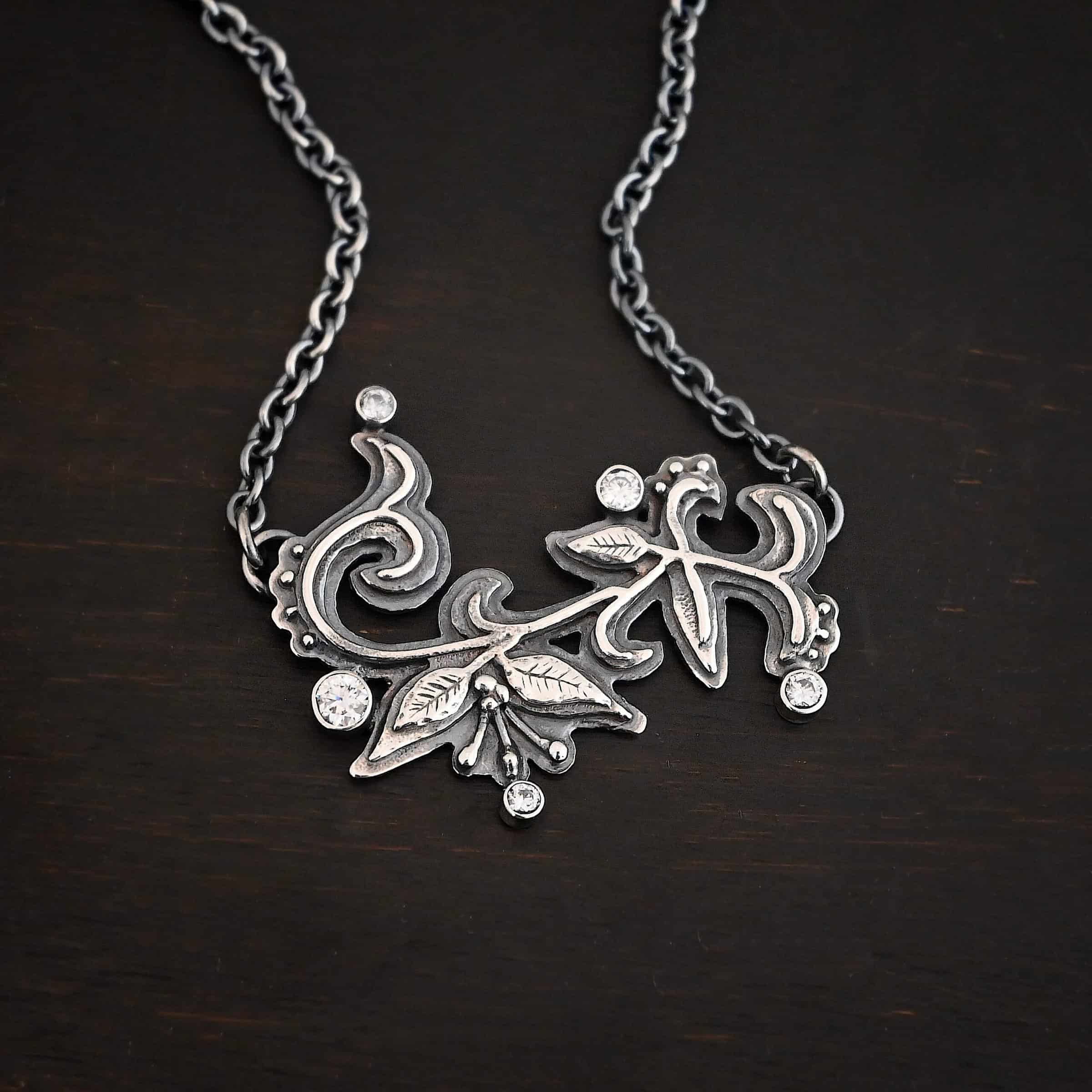White Satin Necklace Photo of the "White Satin" Necklace. Made out of sterling silver and moissanites. There are five moissanites scattered around the outside edge of the pendant. The rest of the pendant is three layers of silver that flow from the left to the right. Attached around the swirl are three silver leaves, along with silver dots and small lines of silver with silver balls on the end of them. Each side of the pendant has a loop soldered to the back of it. The chain of the necklace is attached to the loops on the back. Necklace is lying against a dark background.