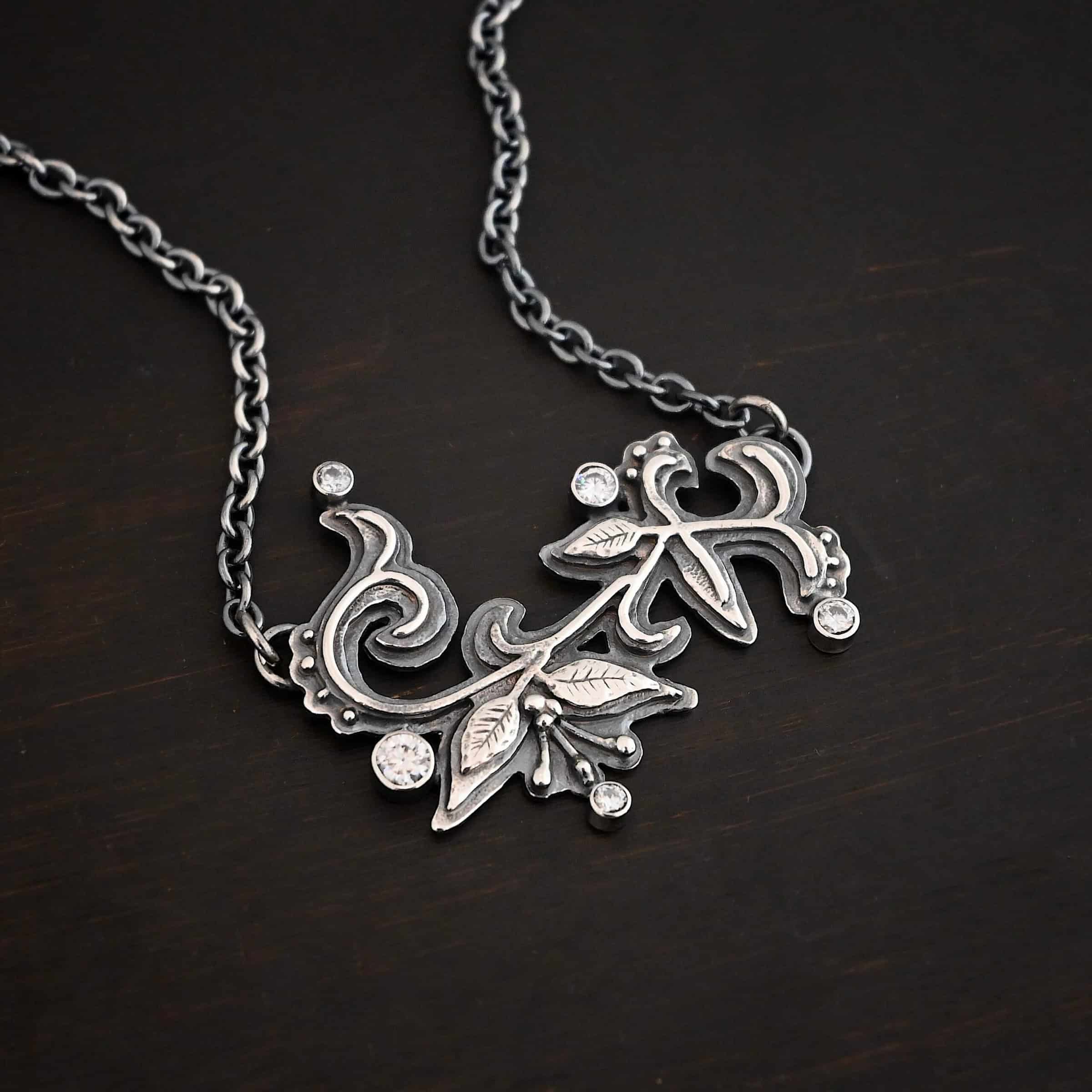 White Satin Necklace Photo of the "White Satin" Necklace. Made out of sterling silver and moissanites. There are five moissanites scattered around the outside edge of the pendant. The rest of the pendant is three layers of silver that flow from the left to the right. Attached around the swirl are three silver leaves, along with silver dots and small lines of silver with silver balls on the end of them. Each side of the pendant has a loop soldered to the back of it. The chain of the necklace is attached to the loops on the back. Necklace is lying against a dark background.