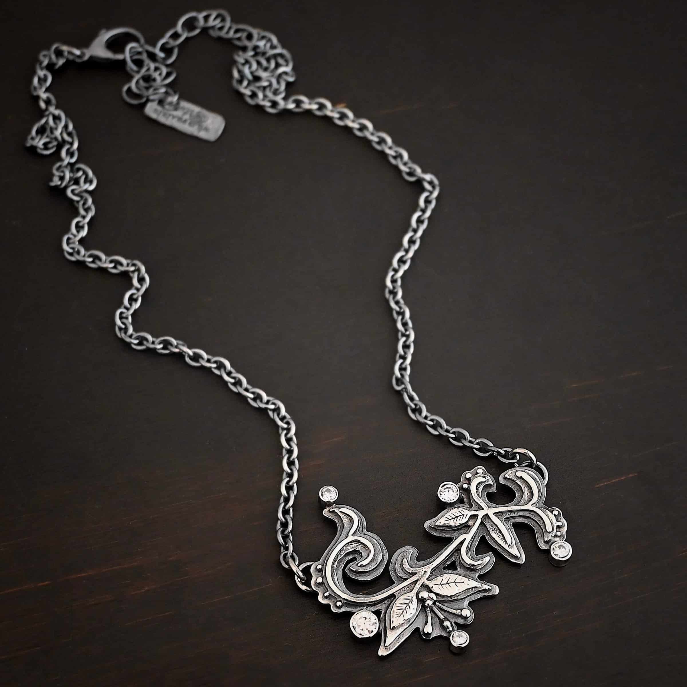 White Satin Necklace Photo of the "White Satin" Necklace. Made out of sterling silver and moissanites. There are five moissanites scattered around the outside edge of the pendant. The rest of the pendant is three layers of silver that flow from the left to the right. Attached around the swirl are three silver leaves, along with silver dots and small lines of silver with silver balls on the end of them. Each side of the pendant has a loop soldered to the back of it. The chain of the necklace is attached to the loops on the back. Necklace is lying against a dark background.