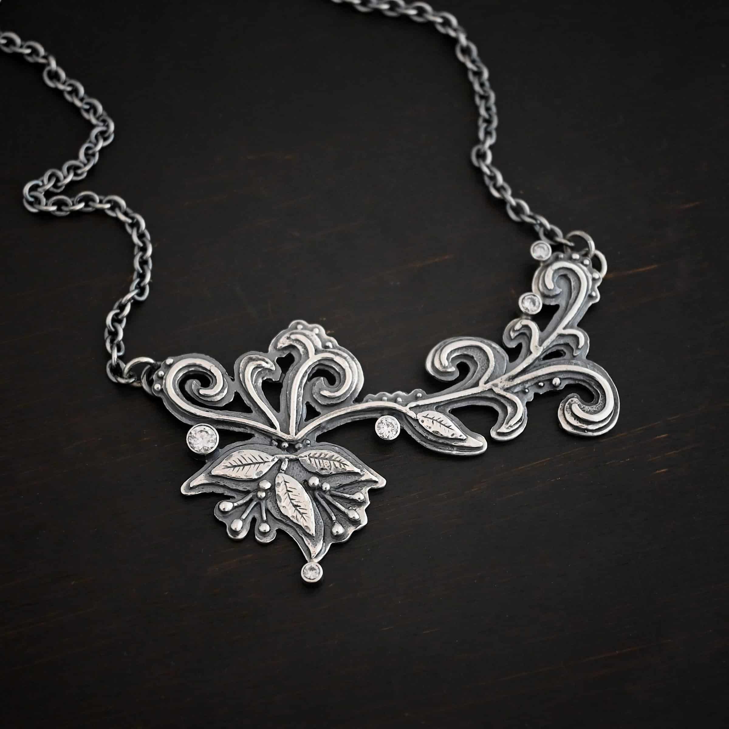 White Veil Necklace Photo of the "White Veil" Necklace. Made out of sterling silver and moissanites. There are five moissanites scattered around the outside edge of the pendant. The rest of the pendant is three layers of silver that flow from the left to the right. Attached around the swirl are three silver leaves, along with silver dots and small lines of silver with silver balls on the end of them. Each side of the pendant has a loop soldered to the back of it. The chain of the necklace is attached to the loops on the back. Necklace is lying against a dark background.