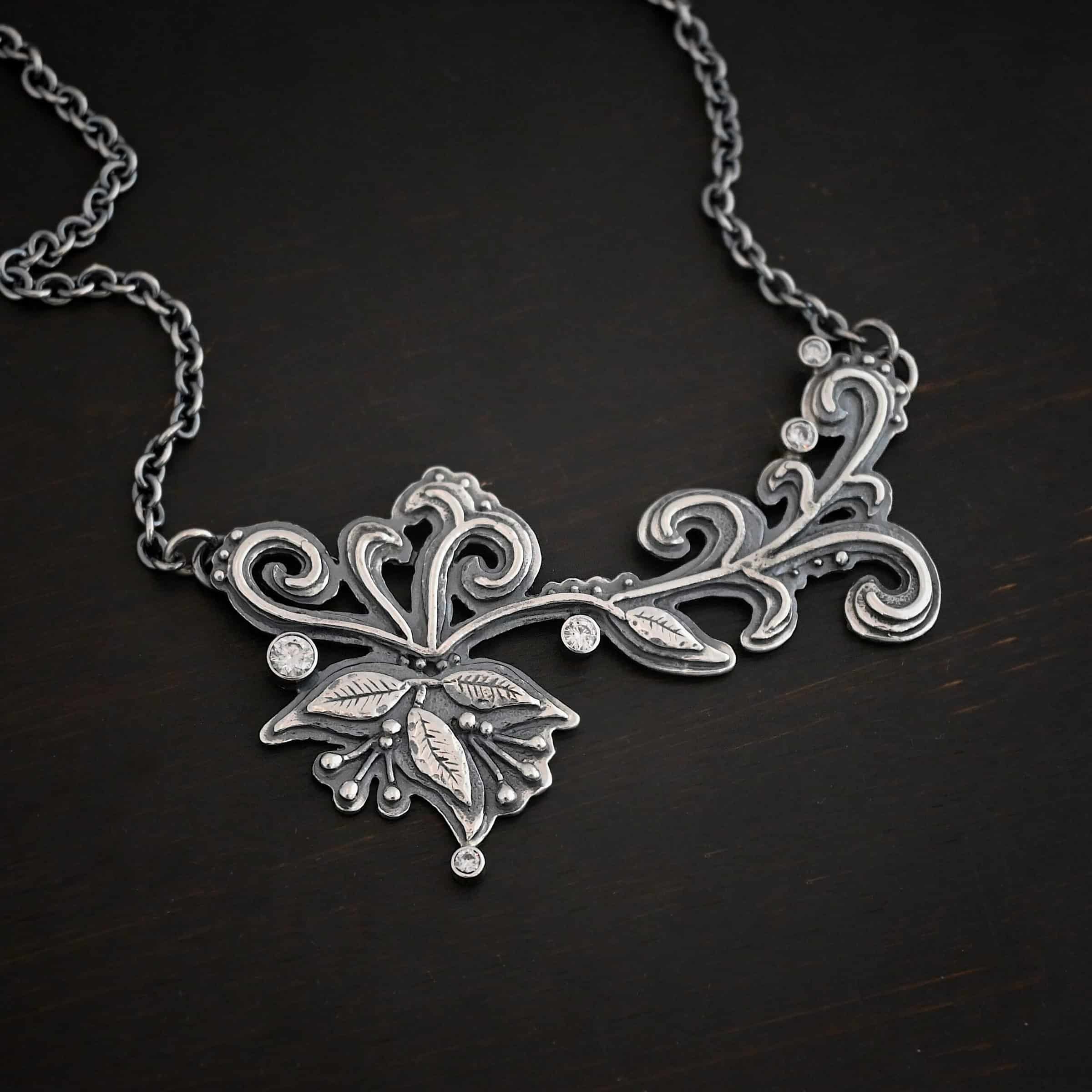 White Veil Necklace Photo of the "White Veil" Necklace. Made out of sterling silver and moissanites. There are five moissanites scattered around the outside edge of the pendant. The rest of the pendant is three layers of silver that flow from the left to the right. Attached around the swirl are three silver leaves, along with silver dots and small lines of silver with silver balls on the end of them. Each side of the pendant has a loop soldered to the back of it. The chain of the necklace is attached to the loops on the back. Necklace is lying against a dark background.