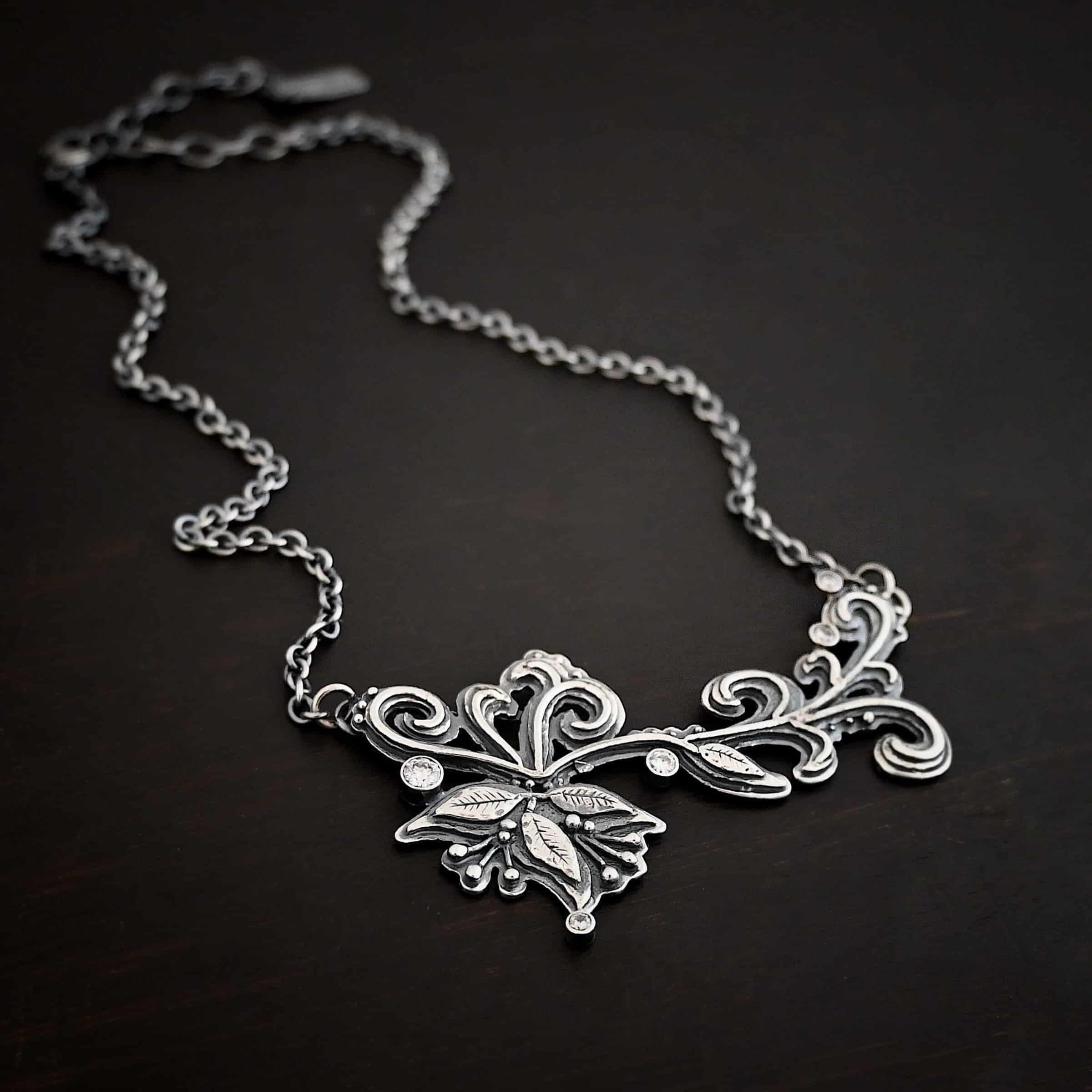 White Veil Necklace Photo of the "White Veil" Necklace. Made out of sterling silver and moissanites. There are five moissanites scattered around the outside edge of the pendant. The rest of the pendant is three layers of silver that flow from the left to the right. Attached around the swirl are three silver leaves, along with silver dots and small lines of silver with silver balls on the end of them. Each side of the pendant has a loop soldered to the back of it. The chain of the necklace is attached to the loops on the back. Necklace is lying against a dark background.