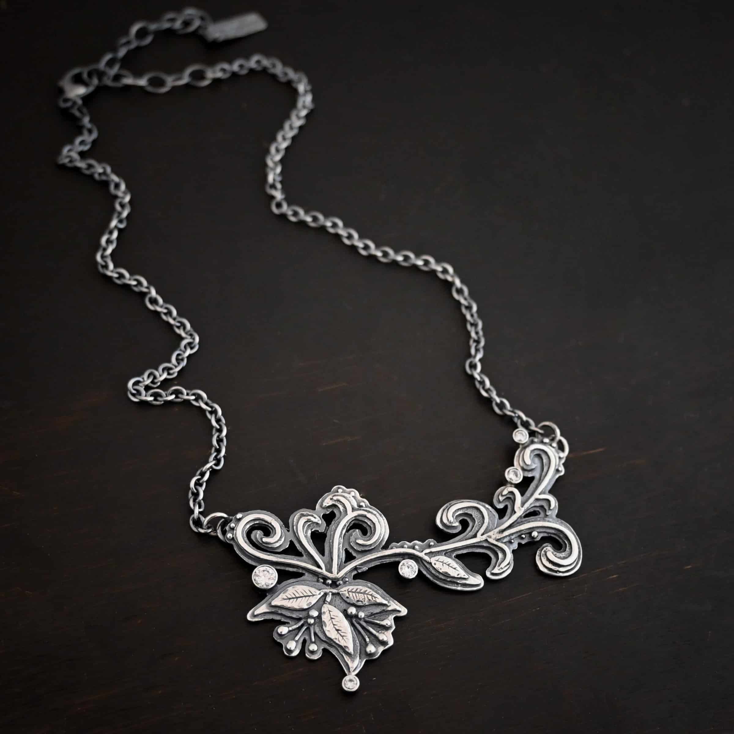 White Veil Necklace Photo of the "White Veil" Necklace. Made out of sterling silver and moissanites. There are five moissanites scattered around the outside edge of the pendant. The rest of the pendant is three layers of silver that flow from the left to the right. Attached around the swirl are three silver leaves, along with silver dots and small lines of silver with silver balls on the end of them. Each side of the pendant has a loop soldered to the back of it. The chain of the necklace is attached to the loops on the back. Necklace is lying against a dark background.