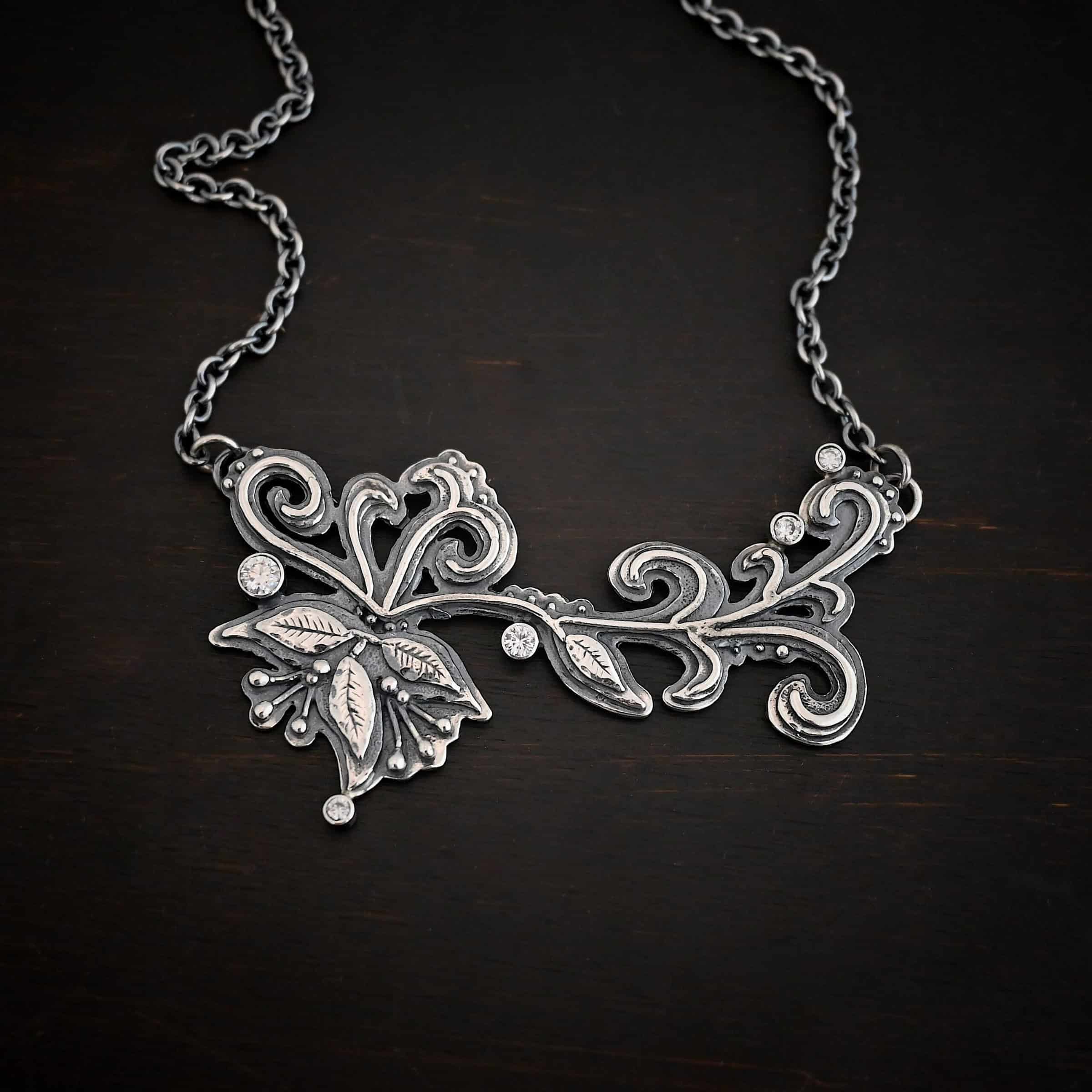 White Veil Necklace Photo of the "White Veil" Necklace. Made out of sterling silver and moissanites. There are five moissanites scattered around the outside edge of the pendant. The rest of the pendant is three layers of silver that flow from the left to the right. Attached around the swirl are three silver leaves, along with silver dots and small lines of silver with silver balls on the end of them. Each side of the pendant has a loop soldered to the back of it. The chain of the necklace is attached to the loops on the back. Necklace is lying against a dark background.