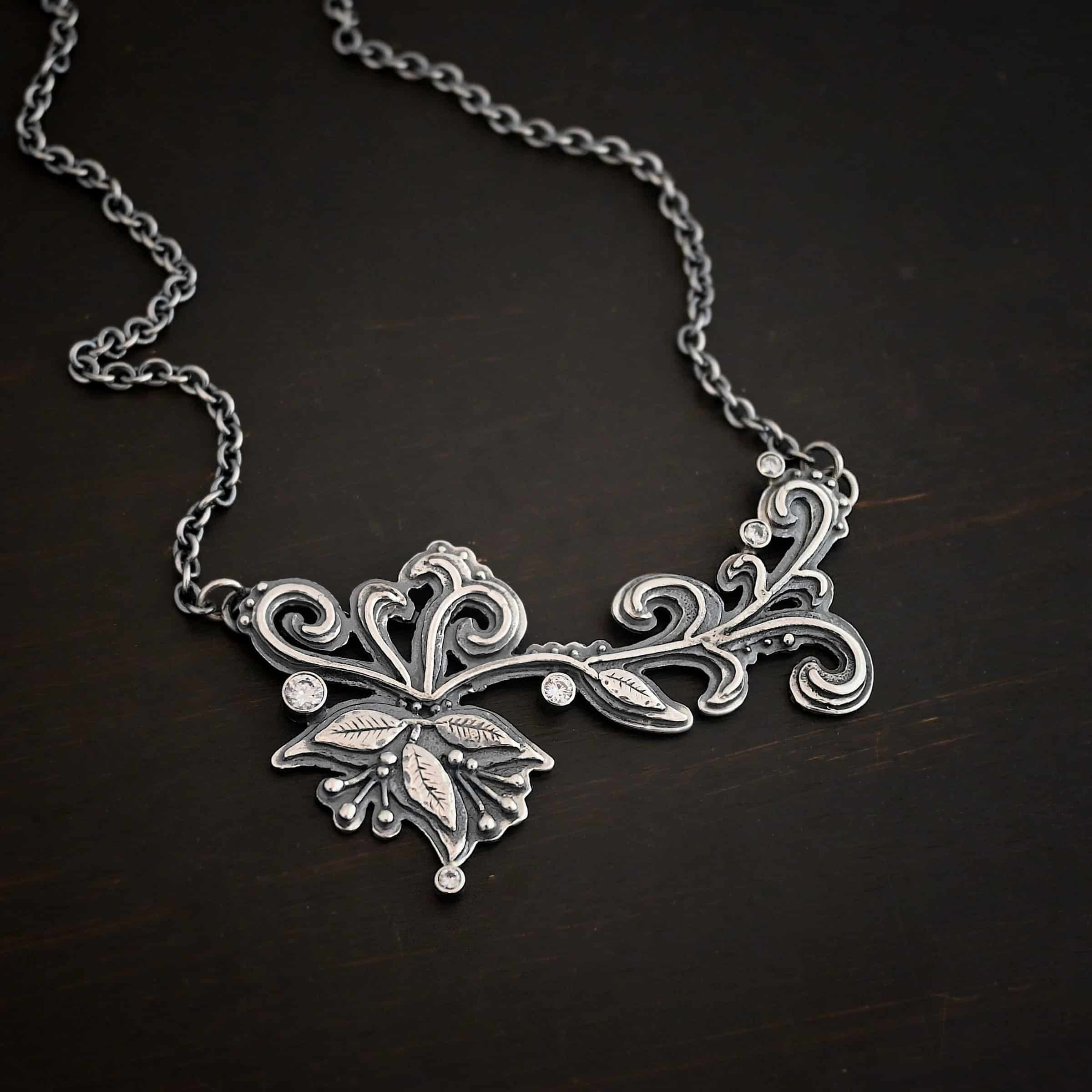 White Veil Necklace Photo of the "White Veil" Necklace. Made out of sterling silver and moissanites. There are five moissanites scattered around the outside edge of the pendant. The rest of the pendant is three layers of silver that flow from the left to the right. Attached around the swirl are three silver leaves, along with silver dots and small lines of silver with silver balls on the end of them. Each side of the pendant has a loop soldered to the back of it. The chain of the necklace is attached to the loops on the back. Necklace is lying against a dark background.