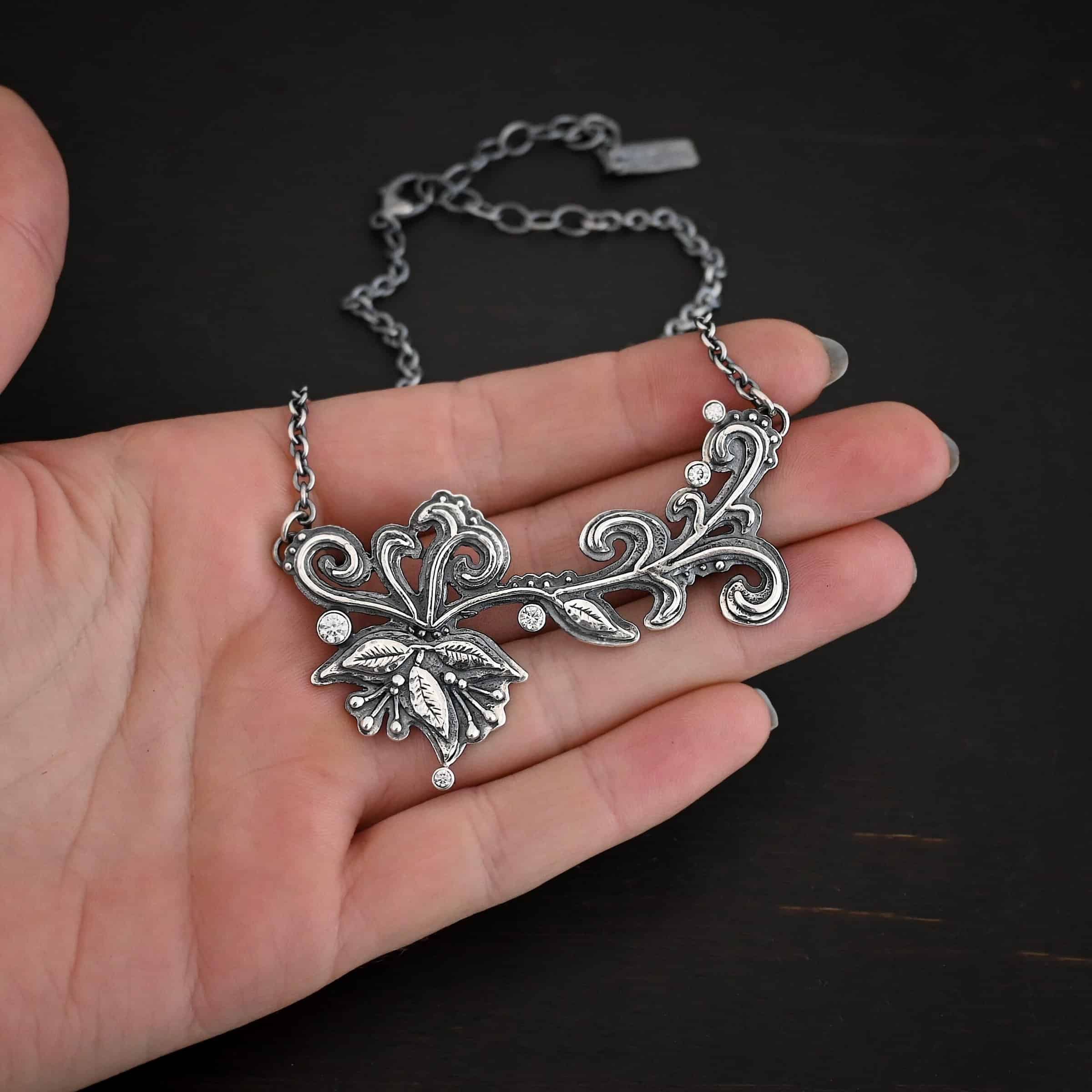 White Veil Necklace Photo of the "White Veil" Necklace. Made out of sterling silver and moissanites. There are five moissanites scattered around the outside edge of the pendant. The rest of the pendant is three layers of silver that flow from the left to the right. Attached around the swirl are three silver leaves, along with silver dots and small lines of silver with silver balls on the end of them. Each side of the pendant has a loop soldered to the back of it. The chain of the necklace is attached to the loops on the back. Necklace is lying against the fingertips of a hand to demonstrate size.