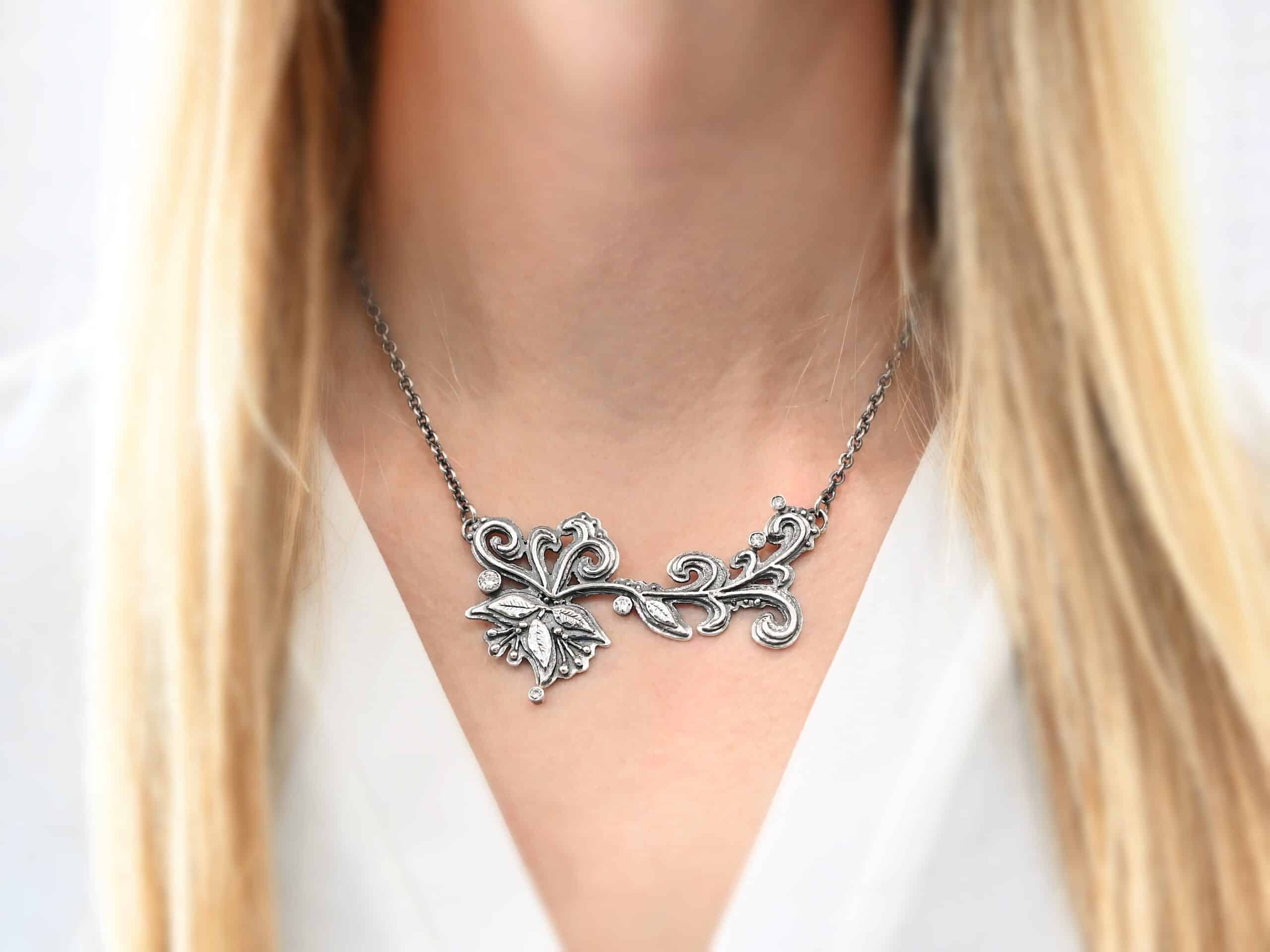 White Veil Necklace Photo of the "White Veil" Necklace. Made out of sterling silver and moissanites. There are five moissanites scattered around the outside edge of the pendant. The rest of the pendant is three layers of silver that flow from the left to the right. Attached around the swirl are three silver leaves, along with silver dots and small lines of silver with silver balls on the end of them. Each side of the pendant has a loop soldered to the back of it. The chain of the necklace is attached to the loops on the back. Necklace is worn by a model to demonstrate size. Model is wearing a white v-neck shirt.