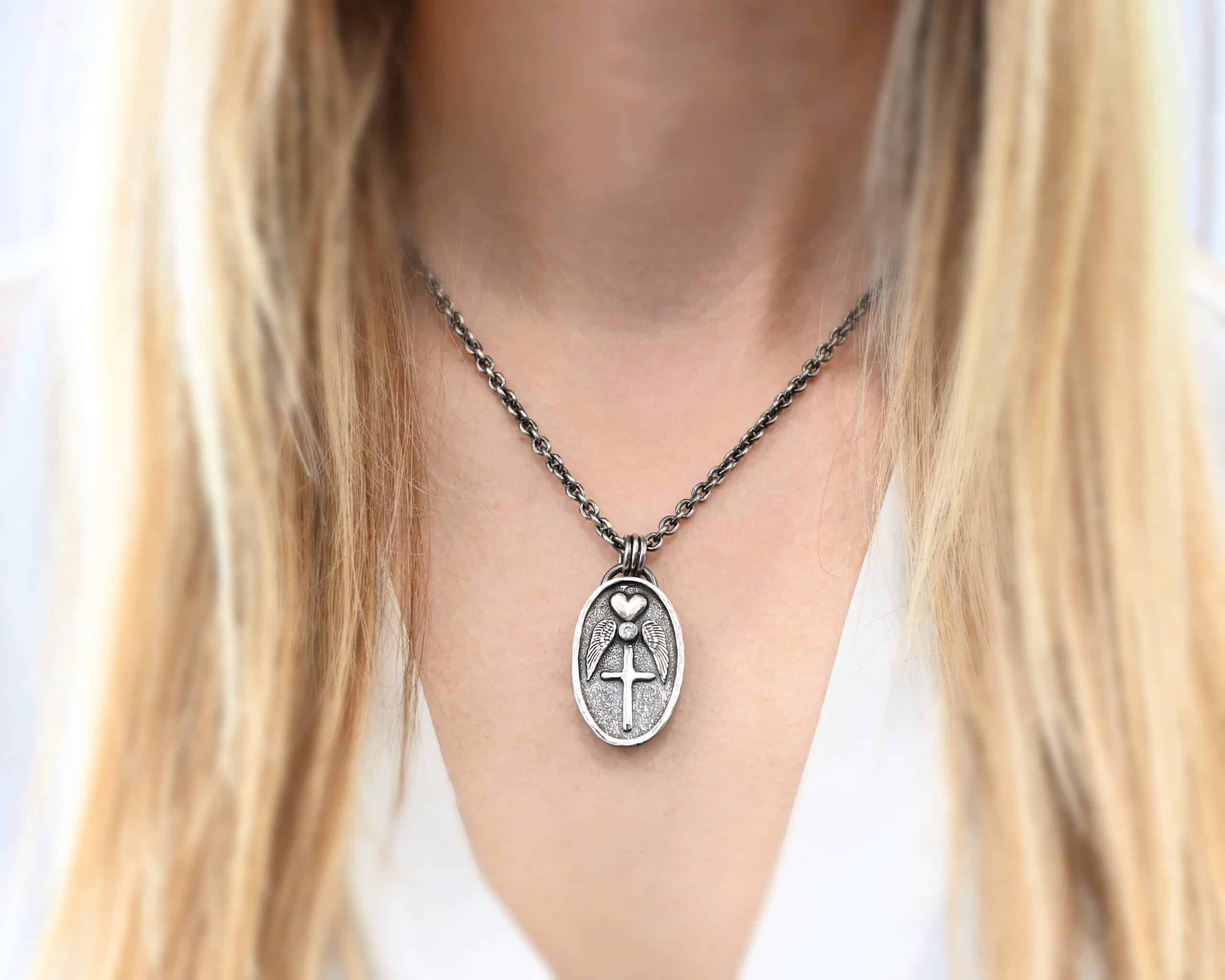 Photo of the "My Forever Angel Cremation Ash" Necklace. Made out of sterling silver. The necklace has a large, oval shaped, thick piece of silver for the pendant. On the inside of the oval is a small heart at the top that sticks up so it is noticeable. There is a small diamond on the bottom tip of the heart. There is an angel wing beneath the heart on each side, surrounding the diamond. Each wing faces outwards. Beneath the ruby is a cross that is soldered. The end of each angel wing touches the left and right side of the cross. There is a thick silver loop soldered to the top. The chain of the necklace goes through the loop. Necklace is worn by a model to demonstrate size.