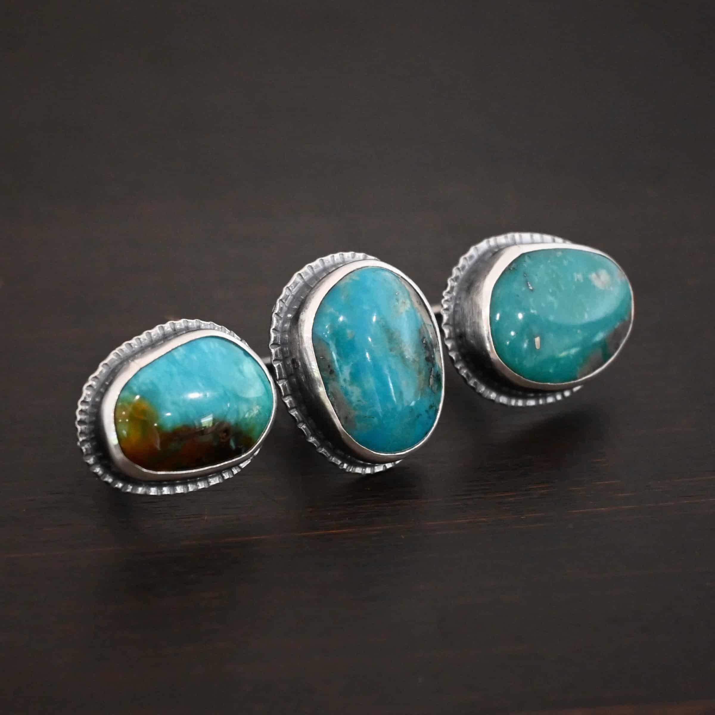 Photo of the "Donna" Ring. Made out of sterling silver and turquoise. There are three oval shaped pieces of turquoise. The ring has two bands that are side by side, so it is worn on two fingers at the same time. The center of the band, where each band meets, has one turquoise that is set vertically. The other two turquoise are set horizontally on the end of each band. Each turquoise stone has hammered silver around it. Ring is sitting against a dark background.