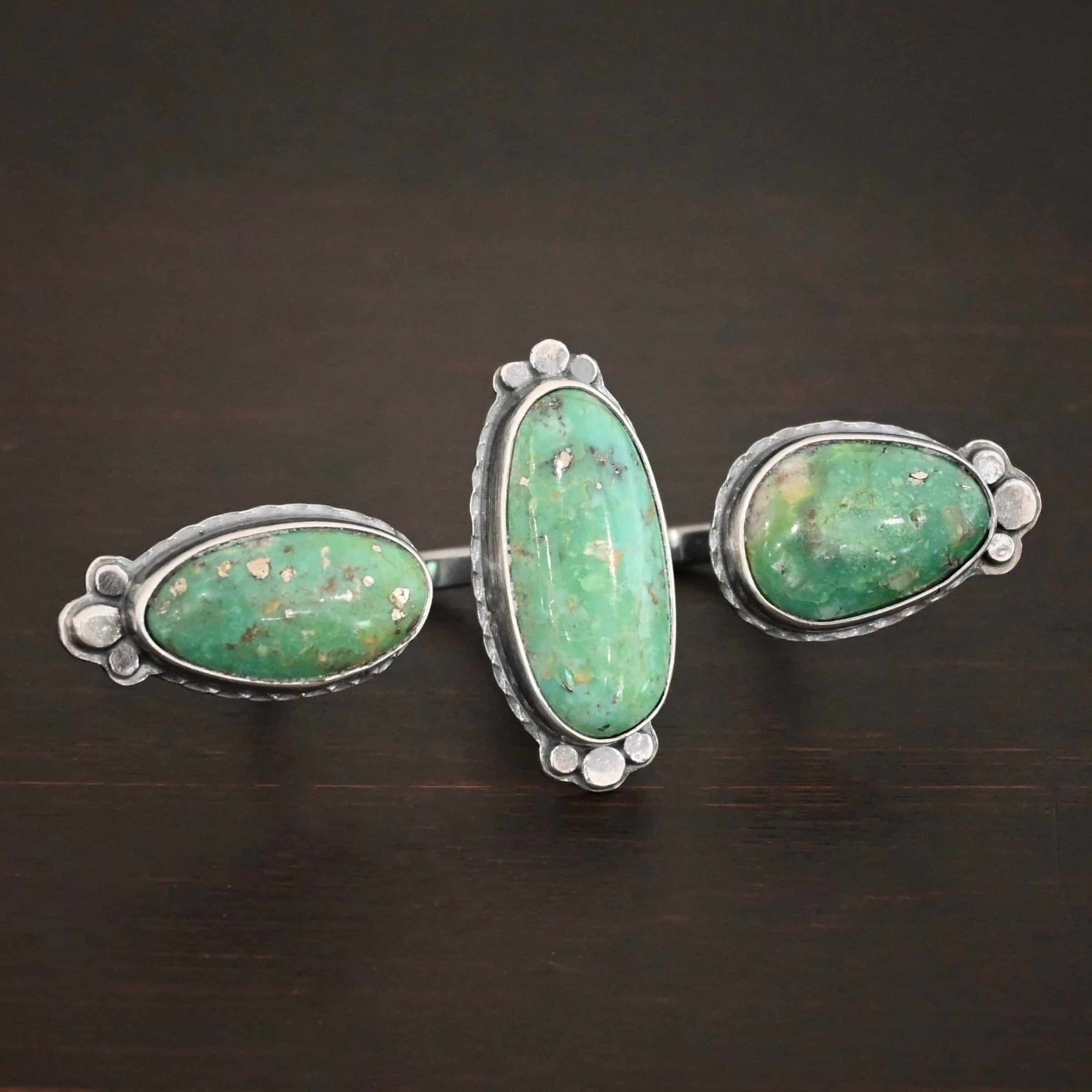 Jenny Ring Photo of the "Jenny" Ring. Made out of sterling silver and turquoise. There are three oval shaped pieces of turquoise. The ring has two bands that are side by side, so it is worn on two fingers at the same time. The center of the band, where each band meets, has one turquoise that is set vertically. The other two turquoise are set horizontally on the end of each band. Each end turquoise has three hammered silver dots that follow the curve of the turquoise on the end. The middle turquoise has six hammered silver dots on it, three on the top and three on the bottom, that follow the curve of the oval. Ring is sitting against a dark background.