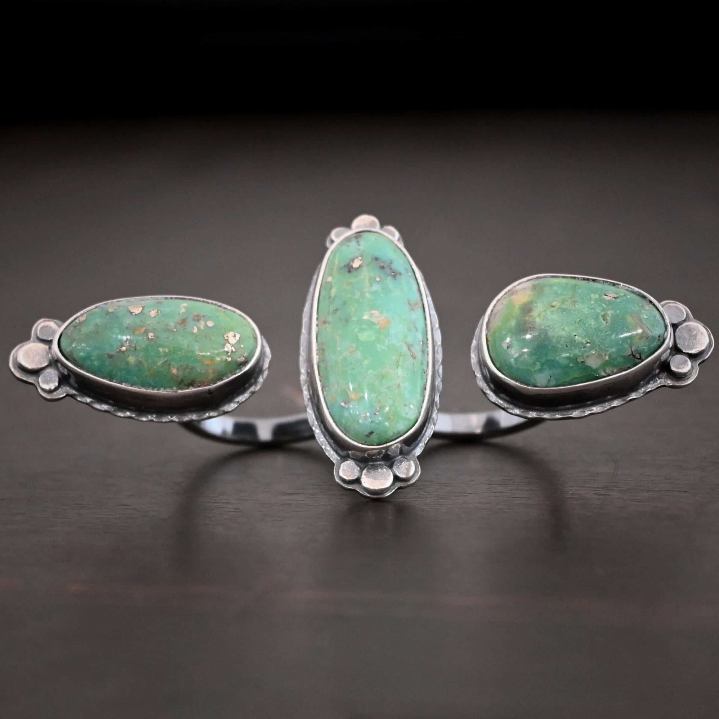 Jenny Ring Photo of the "Jenny" Ring. Made out of sterling silver and turquoise. There are three oval shaped pieces of turquoise. The ring has two bands that are side by side, so it is worn on two fingers at the same time. The center of the band, where each band meets, has one turquoise that is set vertically. The other two turquoise are set horizontally on the end of each band. Each end turquoise has three hammered silver dots that follow the curve of the turquoise on the end. The middle turquoise has six hammered silver dots on it, three on the top and three on the bottom, that follow the curve of the oval. Ring is sitting against a dark background.