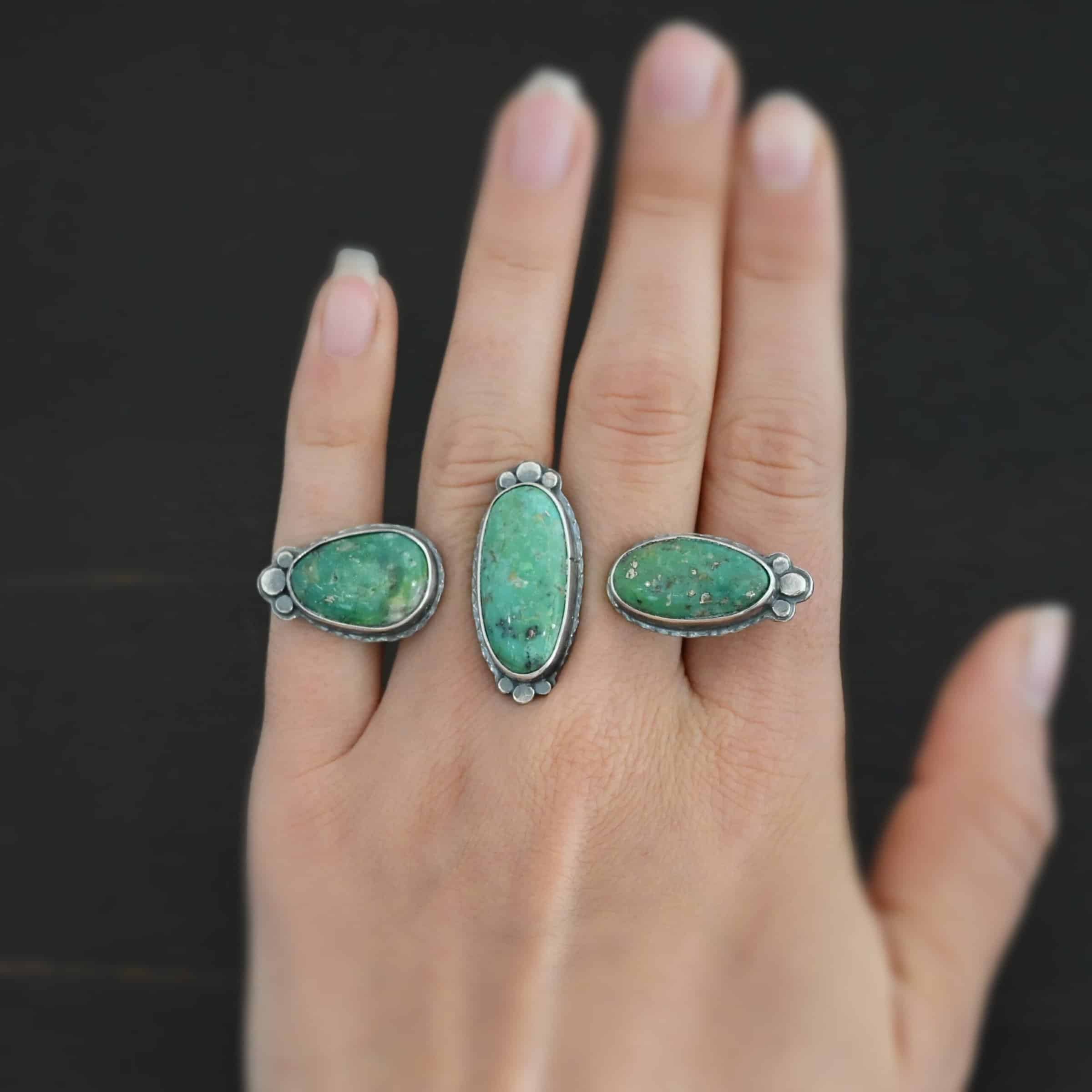 Jenny Ring Photo of the "Jenny" Ring. Made out of sterling silver and turquoise. There are three oval shaped pieces of turquoise. The ring has two bands that are side by side, so it is worn on two fingers at the same time. The center of the band, where each band meets, has one turquoise that is set vertically. The other two turquoise are set horizontally on the end of each band. Each end turquoise has three hammered silver dots that follow the curve of the turquoise on the end. The middle turquoise has six hammered silver dots on it, three on the top and three on the bottom, that follow the curve of the oval. Ring is worn by a model.