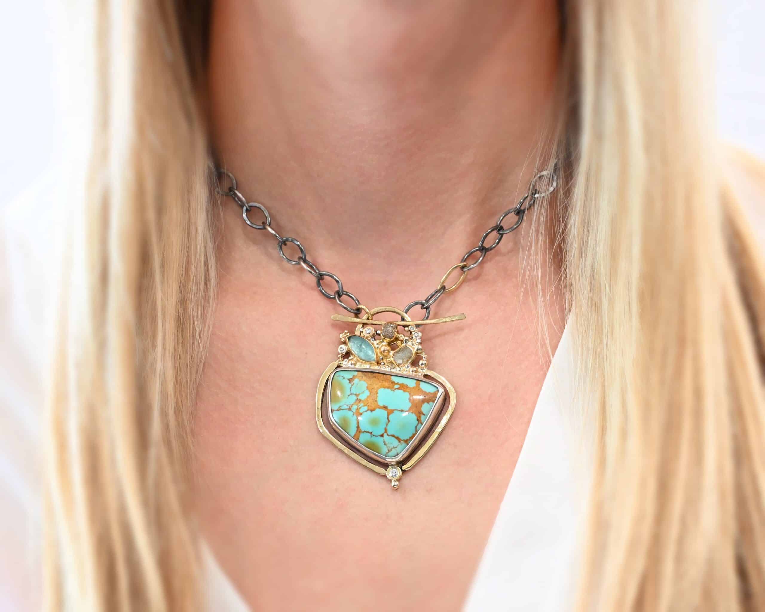 Photo of the "Golden Hour" Necklace. It is made out of diamonds, aquamarine, & kingman turquoise. The turquoise is the largest stone, and it is a unique shape. The top of it and sides resemble a square, but the bottom resembles a triangle. On the top of the turquoise is the aquamarine and diamond stones. There are three diamonds and one aquamarine, all set in gold. There is a diamond on the bottom of the turquoise. There is gold going around the turquoise and leading to the top where the rest of the stone are. The back of the necklace has a gold loop with a gold bar going through it to create a part of the clasp of the necklace. The chain of the necklace is silver, but there is one gold link. The clasp on the back of the necklace is gold, and there is a gold cross and angel wing charm on the back as well. Necklace is worn by a model at 16 inches long to demonstrate size. Model is wearing a white v-neck shirt.