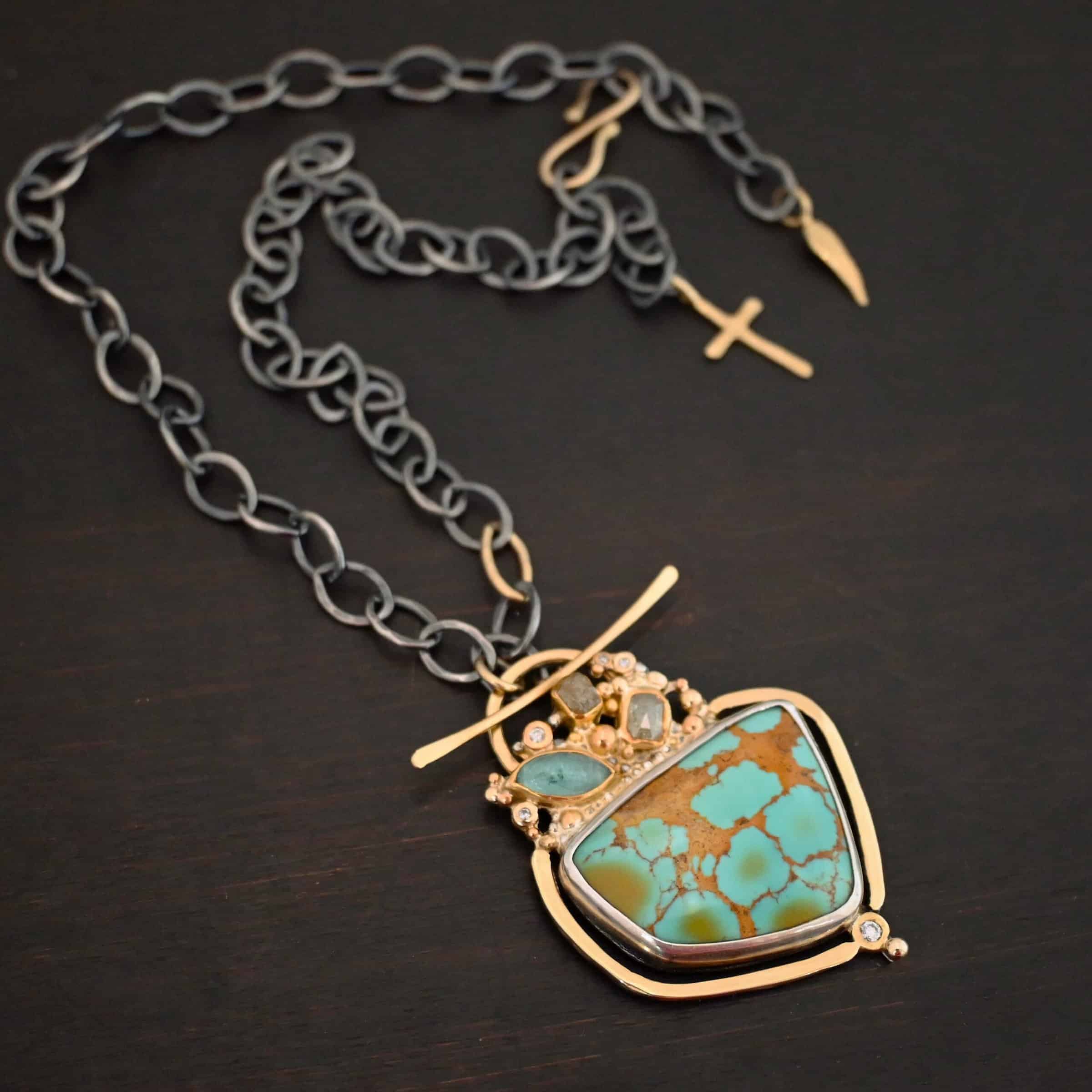 Photo of the "Golden Hour" Necklace. It is made out of diamonds, aquamarine, & kingman turquoise. The turquoise is the largest stone, and it is a unique shape. The top of it and sides resemble a square, but the bottom resembles a triangle. On the top of the turquoise is the aquamarine and diamond stones. There are three diamonds and one aquamarine, all set in gold. There is a diamond on the bottom of the turquoise. There is gold going around the turquoise and leading to the top where the rest of the stone are. The back of the necklace has a gold loop with a gold bar going through it to create a part of the clasp of the necklace. The chain of the necklace is silver, but there is one gold link. The clasp on the back of the necklace is gold, and there is a gold cross and angel wing charm on the back as well. Necklace is lying against a dark background.