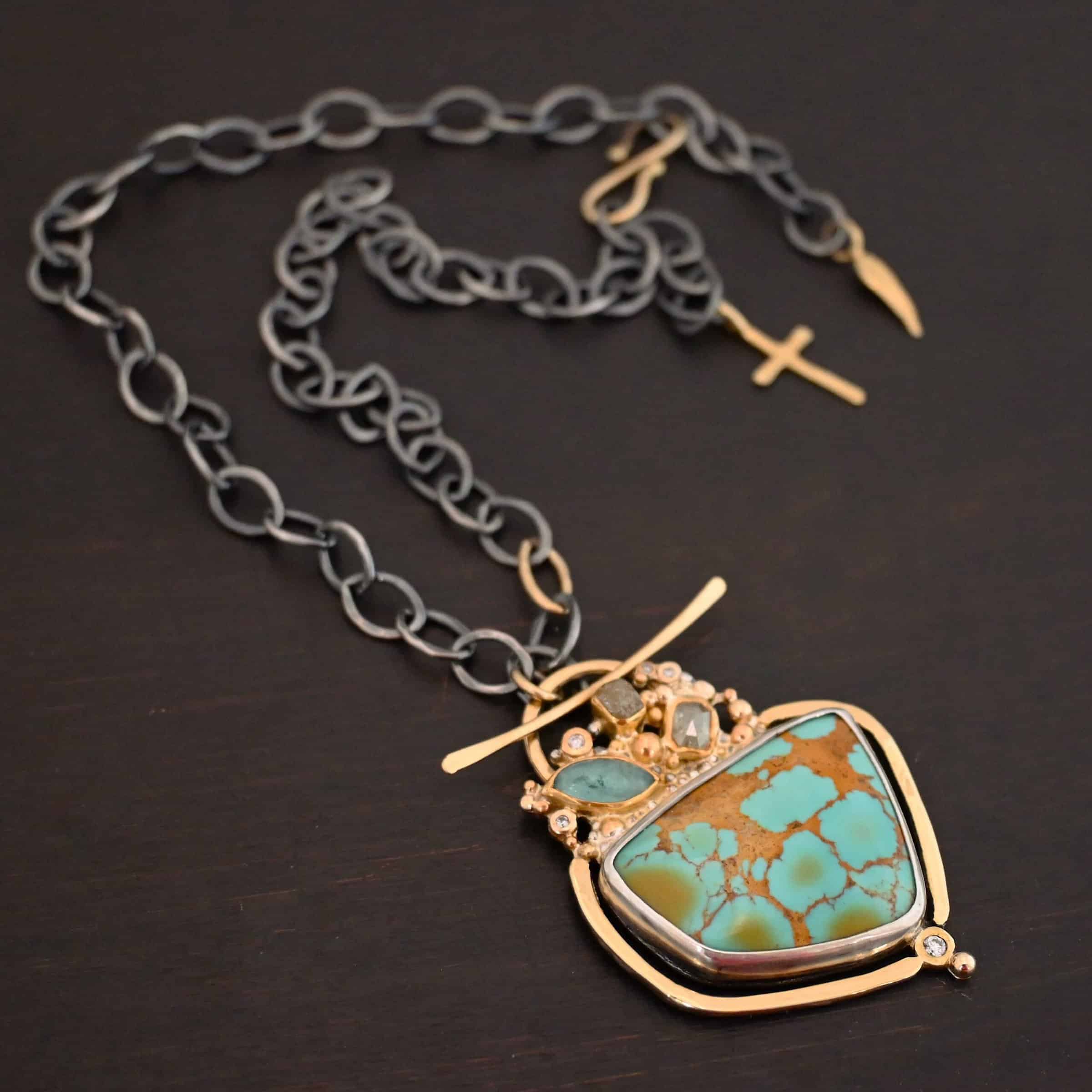 Photo of the "Golden Hour" Necklace. It is made out of diamonds, aquamarine, & kingman turquoise. The turquoise is the largest stone, and it is a unique shape. The top of it and sides resemble a square, but the bottom resembles a triangle. On the top of the turquoise is the aquamarine and diamond stones. There are three diamonds and one aquamarine, all set in gold. There is a diamond on the bottom of the turquoise. There is gold going around the turquoise and leading to the top where the rest of the stone are. The back of the necklace has a gold loop with a gold bar going through it to create a part of the clasp of the necklace. The chain of the necklace is silver, but there is one gold link. The clasp on the back of the necklace is gold, and there is a gold cross and angel wing charm on the back as well. Necklace is lying against a dark background.