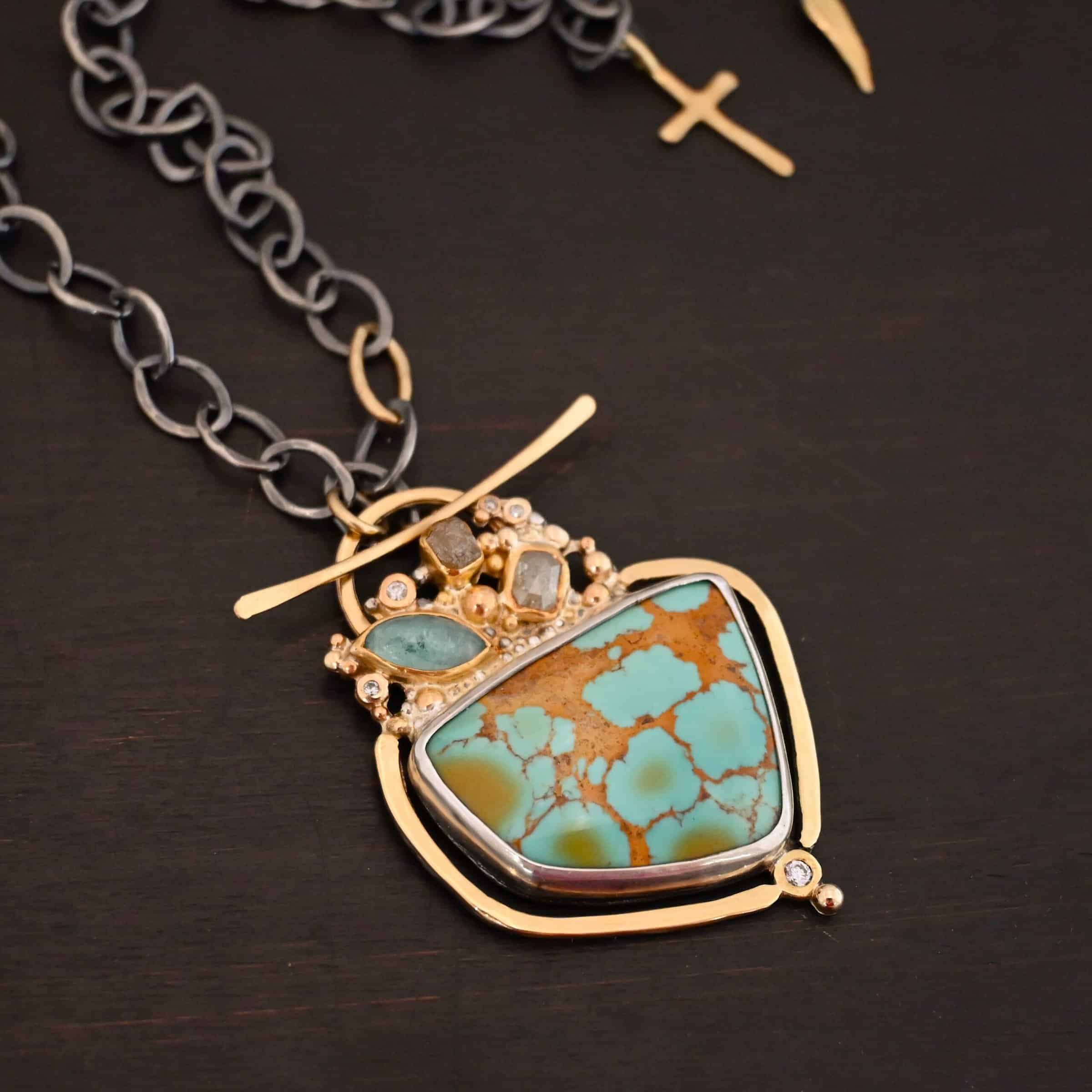 Photo of the "Golden Hour" Necklace. It is made out of diamonds, aquamarine, & kingman turquoise. The turquoise is the largest stone, and it is a unique shape. The top of it and sides resemble a square, but the bottom resembles a triangle. On the top of the turquoise is the aquamarine and diamond stones. There are three diamonds and one aquamarine, all set in gold. There is a diamond on the bottom of the turquoise. There is gold going around the turquoise and leading to the top where the rest of the stone are. The back of the necklace has a gold loop with a gold bar going through it to create a part of the clasp of the necklace. The chain of the necklace is silver, but there is one gold link. The clasp on the back of the necklace is gold, and there is a gold cross and angel wing charm on the back as well. Necklace is lying against a dark background.