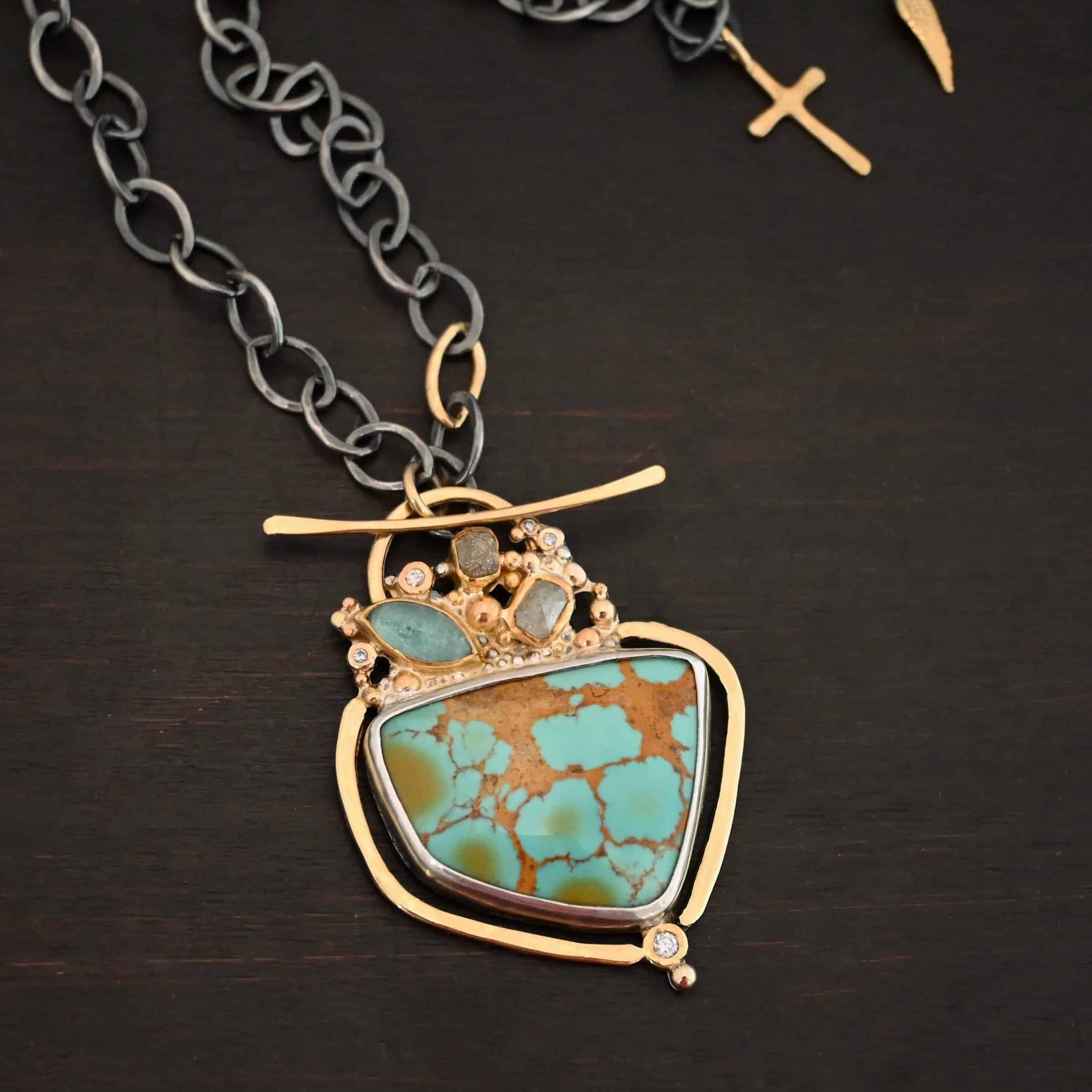 Photo of the "Golden Hour" Necklace. It is made out of diamonds, aquamarine, & kingman turquoise. The turquoise is the largest stone, and it is a unique shape. The top of it and sides resemble a square, but the bottom resembles a triangle. On the top of the turquoise is the aquamarine and diamond stones. There are three diamonds and one aquamarine, all set in gold. There is a diamond on the bottom of the turquoise. There is gold going around the turquoise and leading to the top where the rest of the stone are. The back of the necklace has a gold loop with a gold bar going through it to create a part of the clasp of the necklace. The chain of the necklace is silver, but there is one gold link. The clasp on the back of the necklace is gold, and there is a gold cross and angel wing charm on the back as well. Necklace is lying against a dark background.