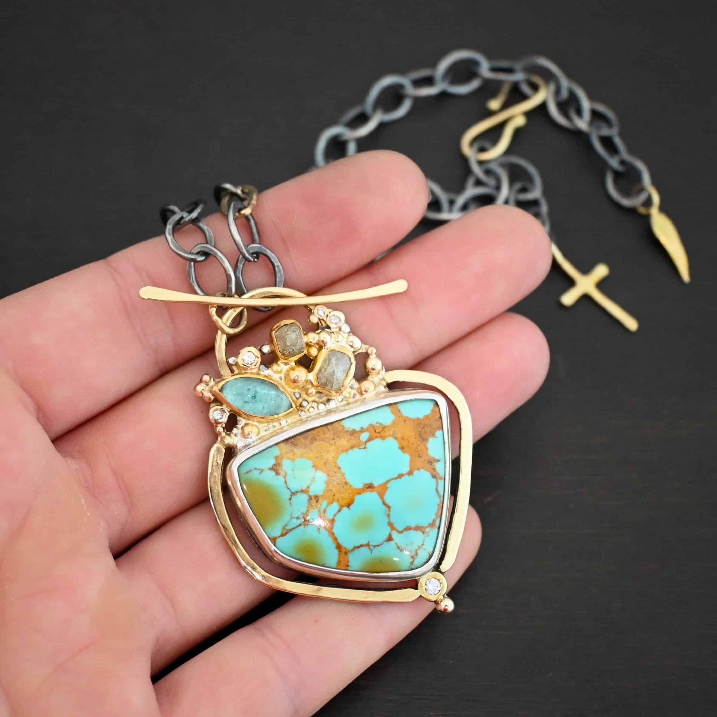 Photo of the "Golden Hour" Necklace. It is made out of diamonds, aquamarine, & kingman turquoise. The turquoise is the largest stone, and it is a unique shape. The top of it and sides resemble a square, but the bottom resembles a triangle. On the top of the turquoise is the aquamarine and diamond stones. There are three diamonds and one aquamarine, all set in gold. There is a diamond on the bottom of the turquoise. There is gold going around the turquoise and leading to the top where the rest of the stone are. The back of the necklace has a gold loop with a gold bar going through it to create a part of the clasp of the necklace. The chain of the necklace is silver, but there is one gold link. The clasp on the back of the necklace is gold, and there is a gold cross and angel wing charm on the back as well. Necklace is lying against the fingertips of a hand to demonstrate size.