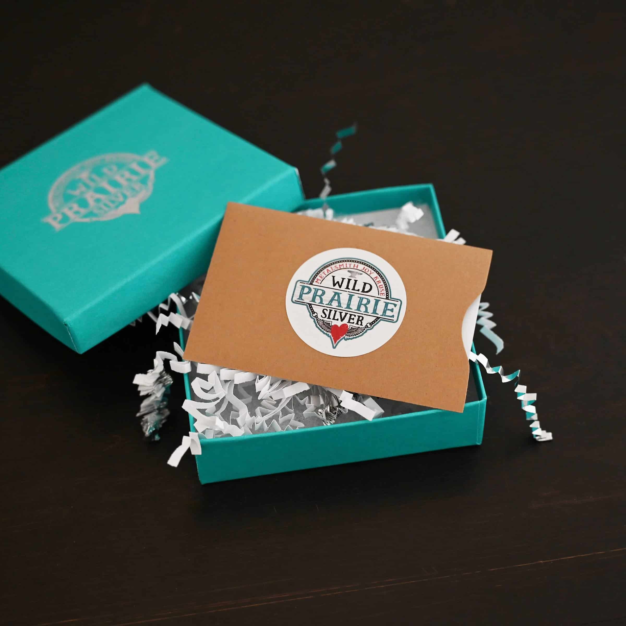 Photo of a "Wild Prairie Silver" gift card. The gift card is white, but it is in a brown paper gift card sleeve/holder. There is a Wild Prairie Silver circle sticker in the center of the gift card holder. The gift card in the holder is lying in a teal box with silver crinkle gift wrap inside of it. The gift wrap is spilling out of the box. The cover for the box is set on the edge of the box on the top left side. Box is set against a dark background.