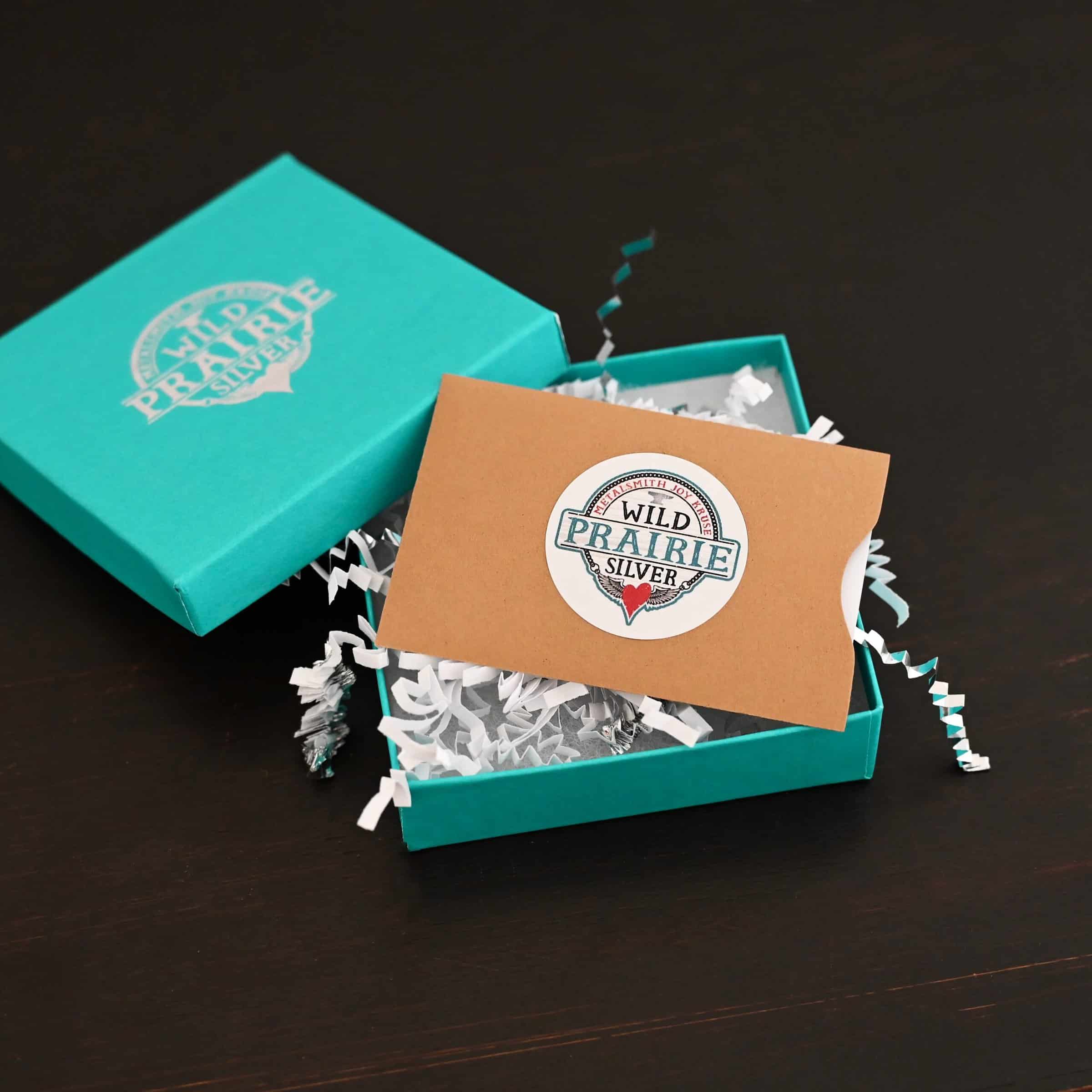 Photo of a "Wild Prairie Silver" gift card. The gift card is white, but it is in a brown paper gift card sleeve/holder. There is a Wild Prairie Silver circle sticker in the center of the gift card holder. The gift card in the holder is lying in a teal box with silver crinkle gift wrap inside of it. The gift wrap is spilling out of the box. The cover for the box is set on the edge of the box on the top left side. Box is set against a dark background.