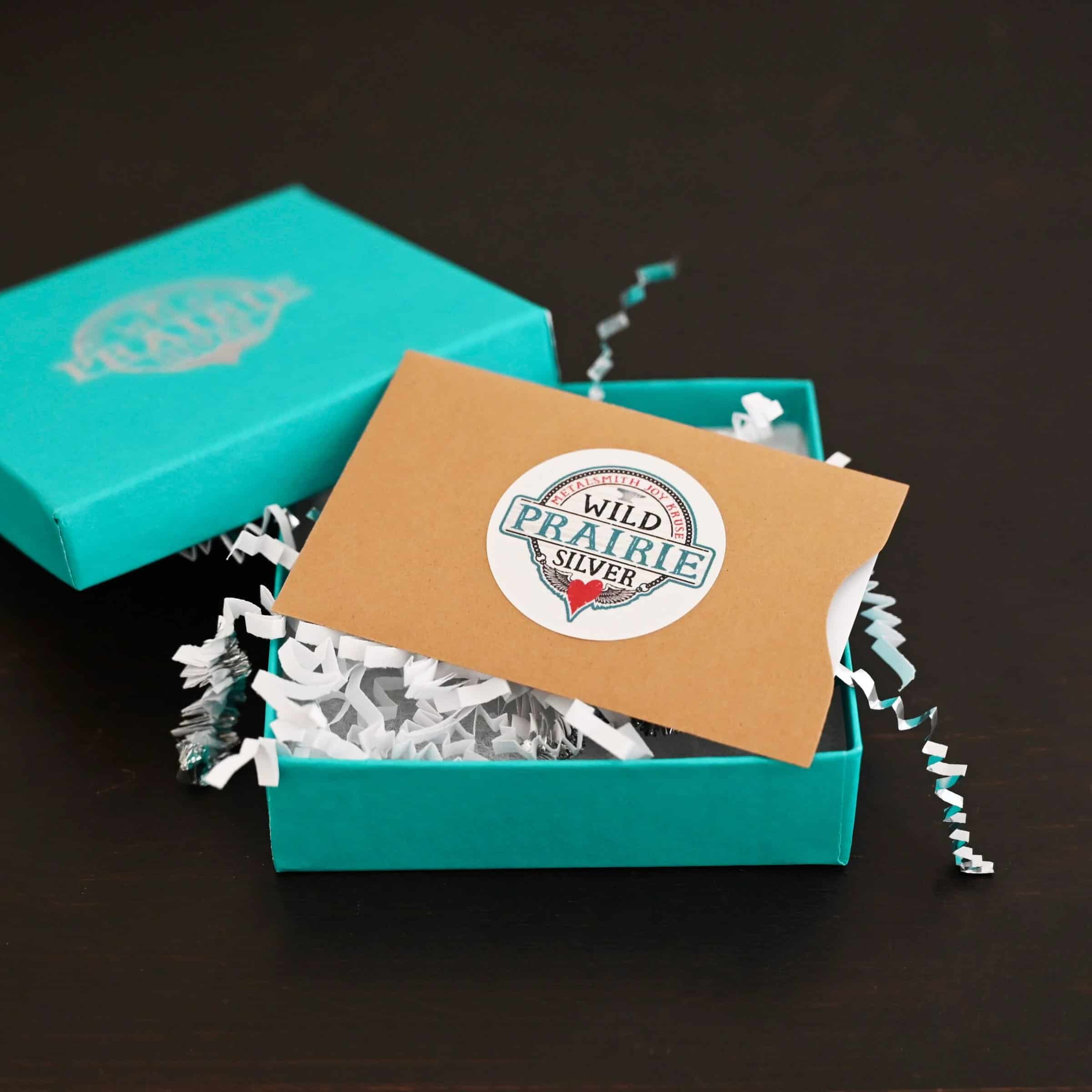 Photo of a "Wild Prairie Silver" gift card. The gift card is white, but it is in a brown paper gift card sleeve/holder. There is a Wild Prairie Silver circle sticker in the center of the gift card holder. The gift card in the holder is lying in a teal box with silver crinkle gift wrap inside of it. The gift wrap is spilling out of the box. The cover for the box is set on the edge of the box on the top left side. Box is set against a dark background.