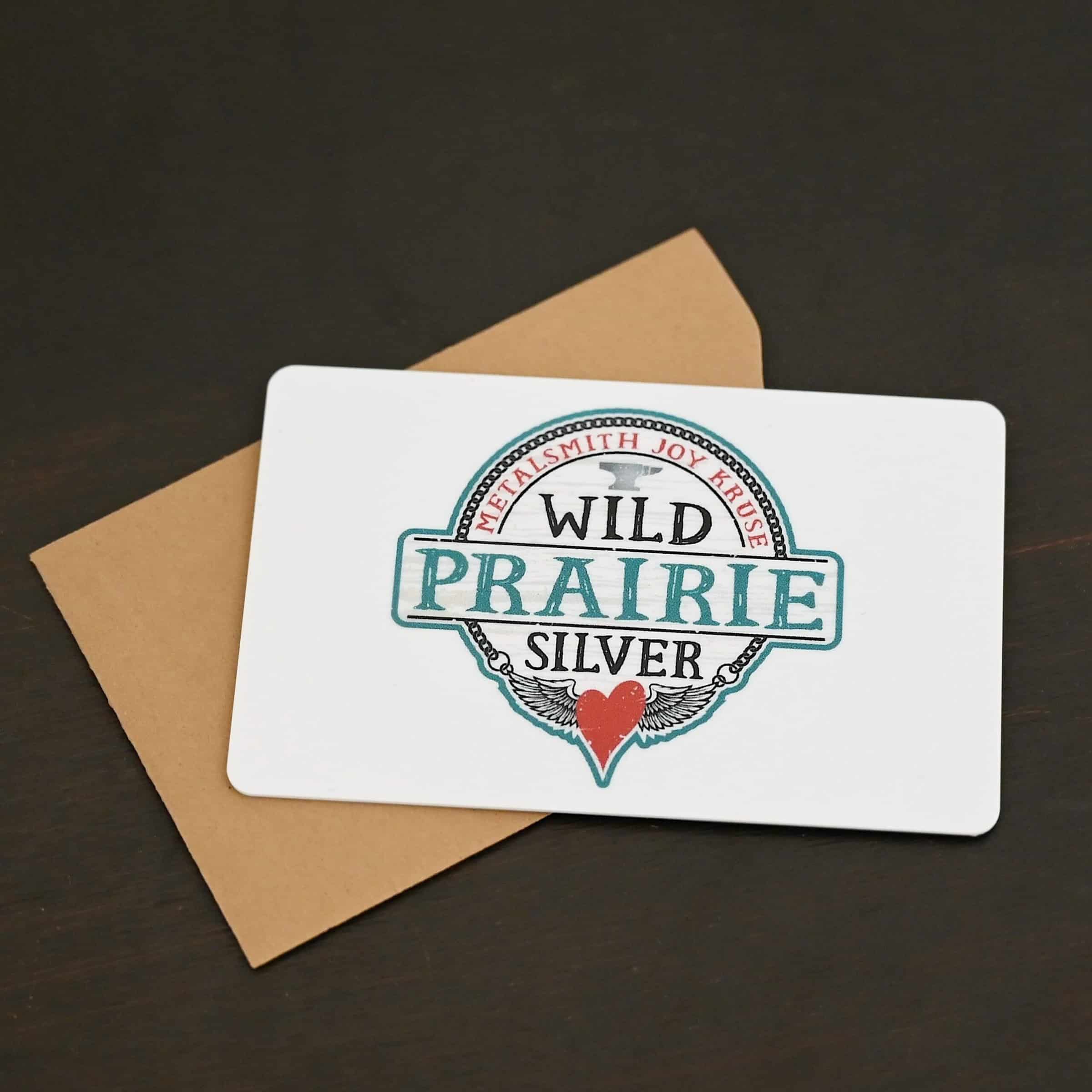 Photo of a "Wild Prairie Silver" gift card. The card is white with a large "Wild Prairie Silver" logo in the center of the card. The logo takes up the entire center of the card. The gift card is lying on a brown paper gift card sleeve/holder. Both the card and gift card holder are lying against a dark background.