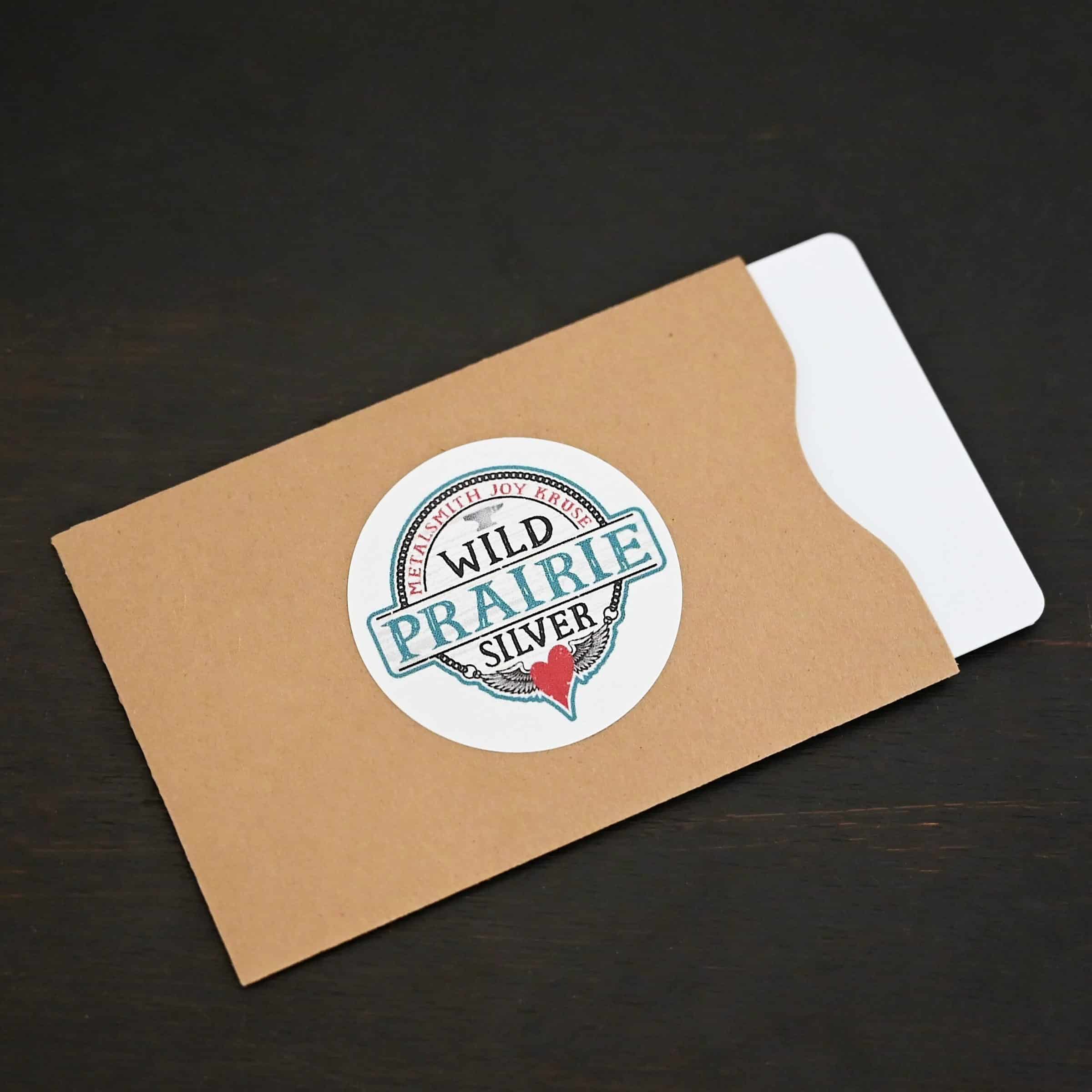 Photo of a "Wild Prairie Silver" gift card. The gift card is white, but it is in a brown paper gift card sleeve/holder. There is a Wild Prairie Silver circle sticker in the center of the gift card holder. The gift card holder is lying against a dark background.