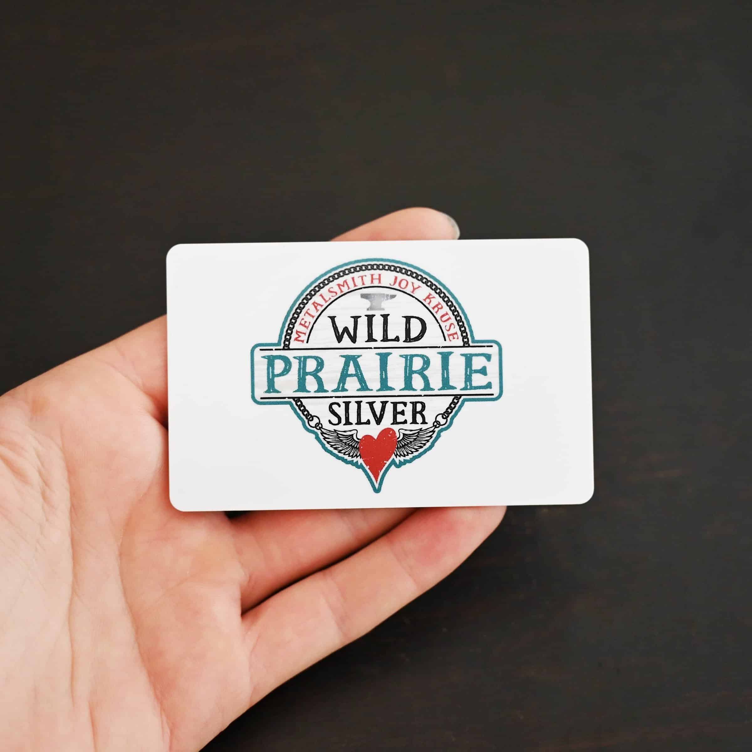 Photo of a "Wild Prairie Silver" gift card. The card is white with a large "Wild Prairie Silver" logo in the center of the card. The logo takes up the entire center of the card. Card is lying in the fingertips of a hand to demonstrate size. Hand is lying against a dark background.