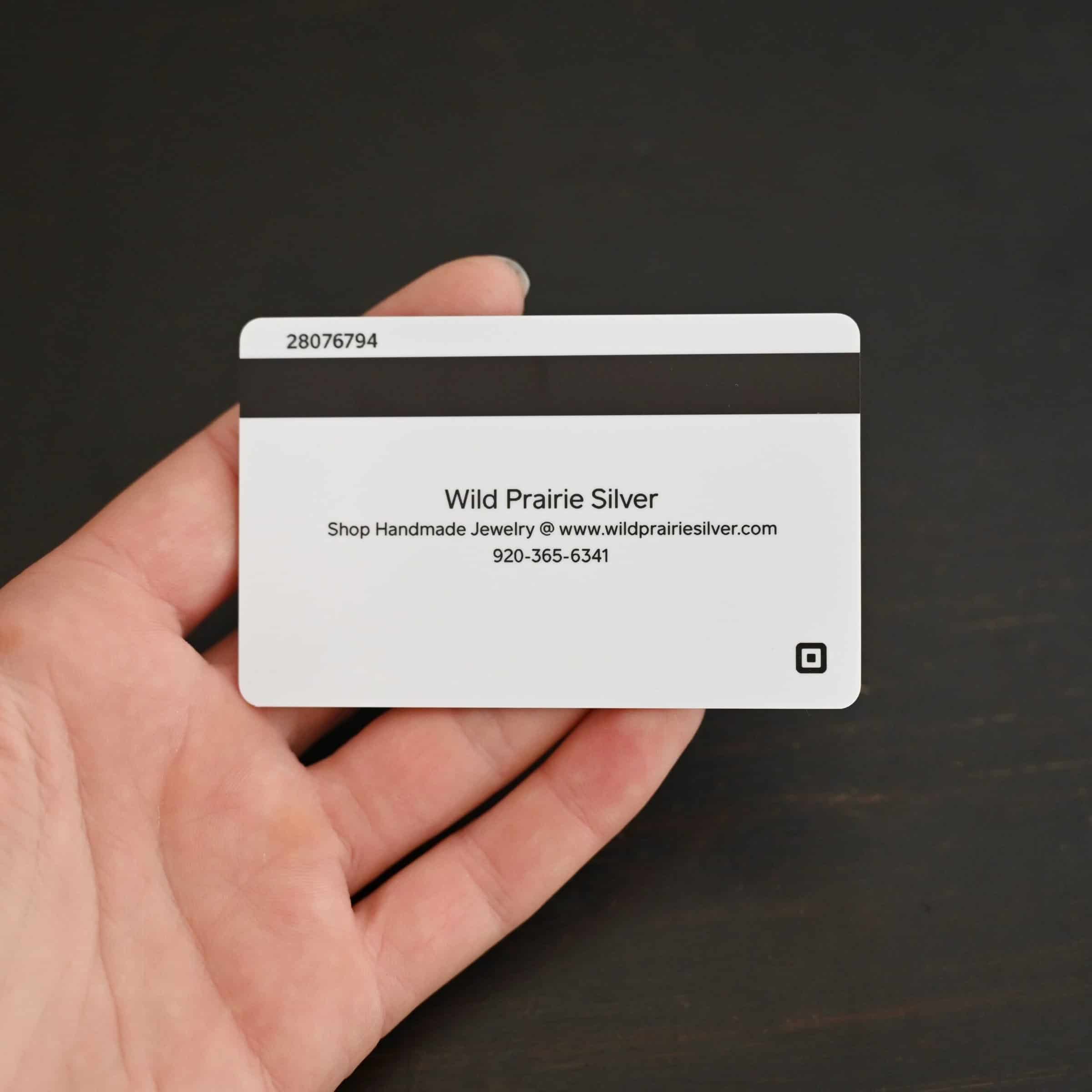 Photo of the back of a "Wild Prairie Silver" gift card. The top numbers say 28076794. Then, there is a thick black line across the top of the card beneath that. Underneath the thick black line are words that say "Wild Prairie Silver" and "Shop Handmade Jewelry @www.wildprairiesilver.com" and "920-365-6341." The square logo is in the bottom right corner of the card. Card is lying on the fingertips of a hand to demonstrate size. Hand is lying against a dark background.