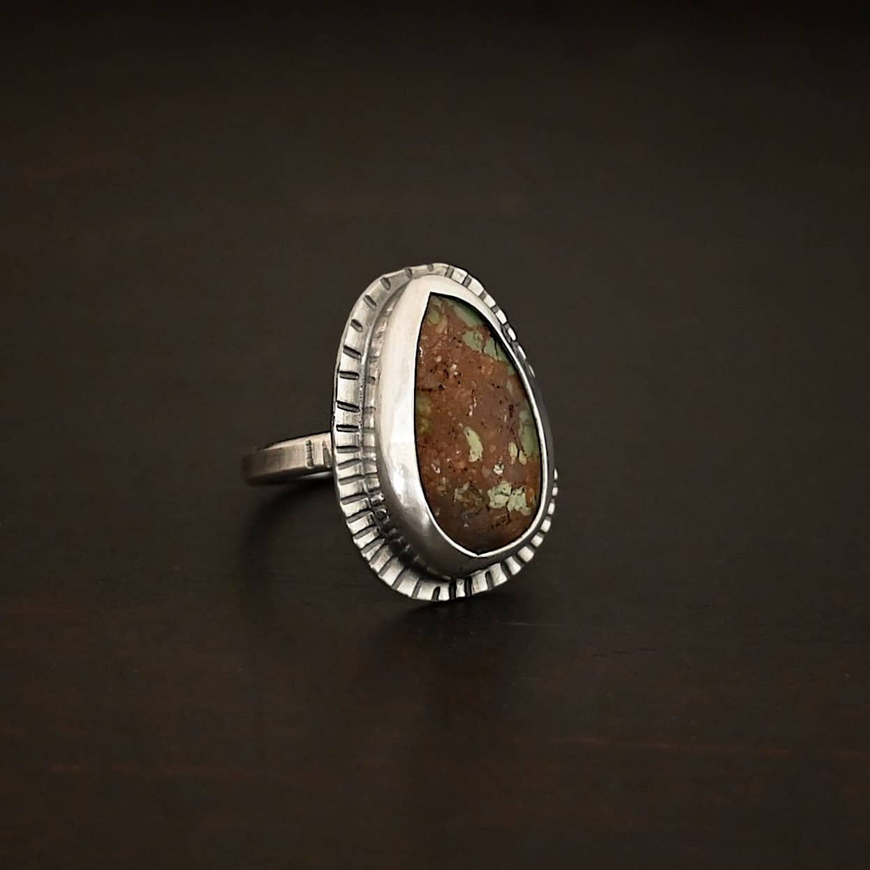 Photo of the "Anthea" Ring. Made out of sterling silver and turquoise. The turquoise is an oval shape. The outside of the ring setting has individual lines hammered all the way around the setting. The lines are darkened to stand out. Ring is sitting against a dark background.