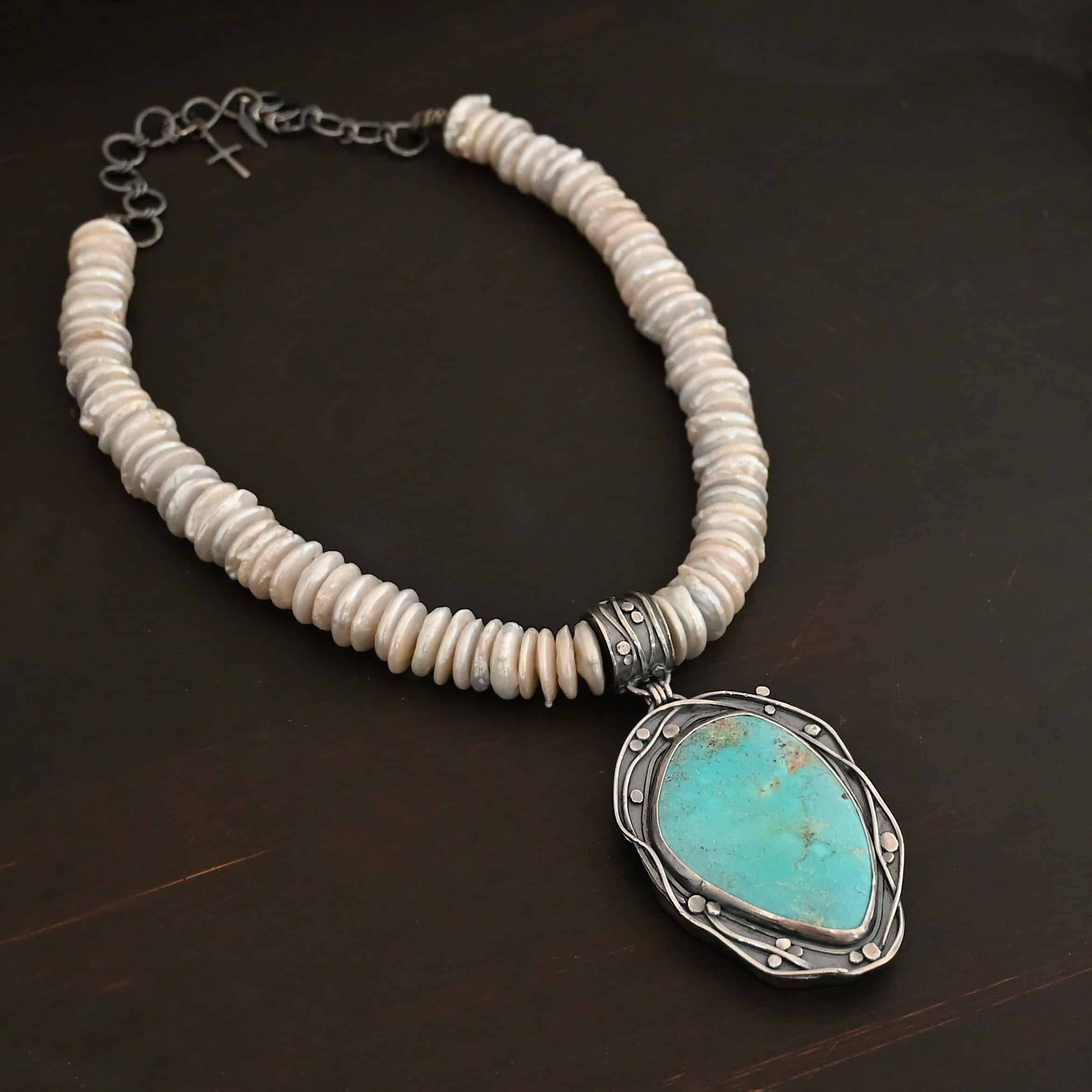 Calypso Necklace Photo of the "Calypso" Necklace. It is a sterling silver, turquoise, and pearl necklace. There is a large, oval shaped turquoise. It is set in sterling silver. The inside of the silver setting has hammered silver dots going all around it, and the outside of the silver setting has swirling silver lines going all around the turquoise. The collar of the necklace is made entirely out of strung pearl disks that are large and rough circle shapes. There is a large silver attachment on the top of the turquoise pendant to attach it to the collar. The attachment has a hole going through the center, and it is in the center of the necklace, with pearls on each side of it. Necklace is laying against a dark background.