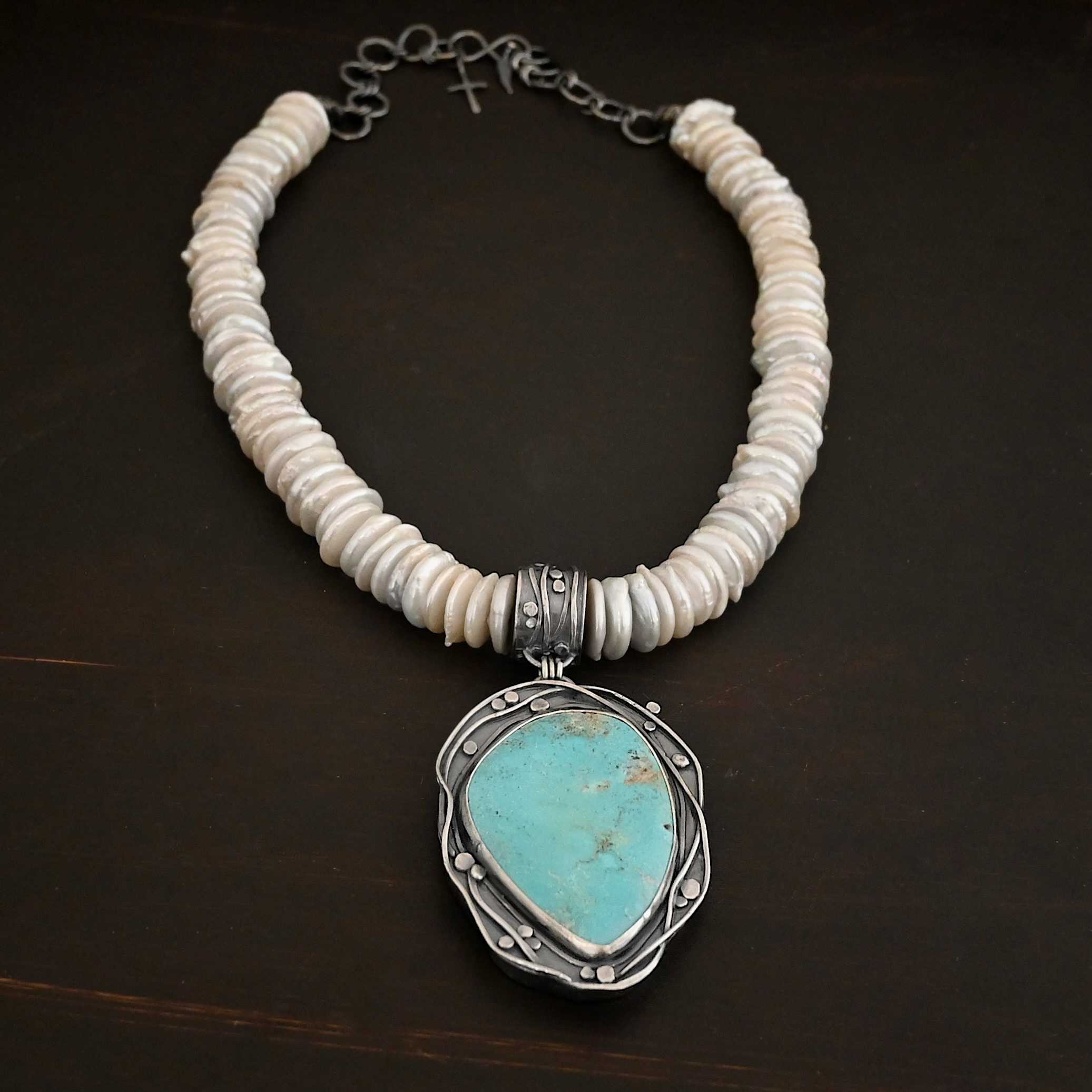 Calypso Necklace Photo of the "Calypso" Necklace. It is a sterling silver, turquoise, and pearl necklace. There is a large, oval shaped turquoise. It is set in sterling silver. The inside of the silver setting has hammered silver dots going all around it, and the outside of the silver setting has swirling silver lines going all around the turquoise. The collar of the necklace is made entirely out of strung pearl disks that are large and rough circle shapes. There is a large silver attachment on the top of the turquoise pendant to attach it to the collar. The attachment has a hole going through the center, and it is in the center of the necklace, with pearls on each side of it. Necklace is laying against a dark background.