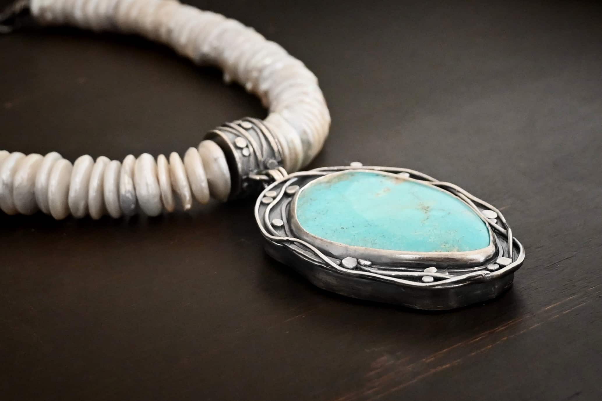 Calypso Necklace Photo of the "Calypso" Necklace. It is a sterling silver, turquoise, and pearl necklace. There is a large, oval shaped turquoise. It is set in sterling silver. The inside of the silver setting has hammered silver dots going all around it, and the outside of the silver setting has swirling silver lines going all around the turquoise. The collar of the necklace is made entirely out of strung pearl disks that are large and rough circle shapes. There is a large silver attachment on the top of the turquoise pendant to attach it to the collar. The attachment has a hole going through the center, and it is in the center of the necklace, with pearls on each side of it. Necklace is laying against a dark background.