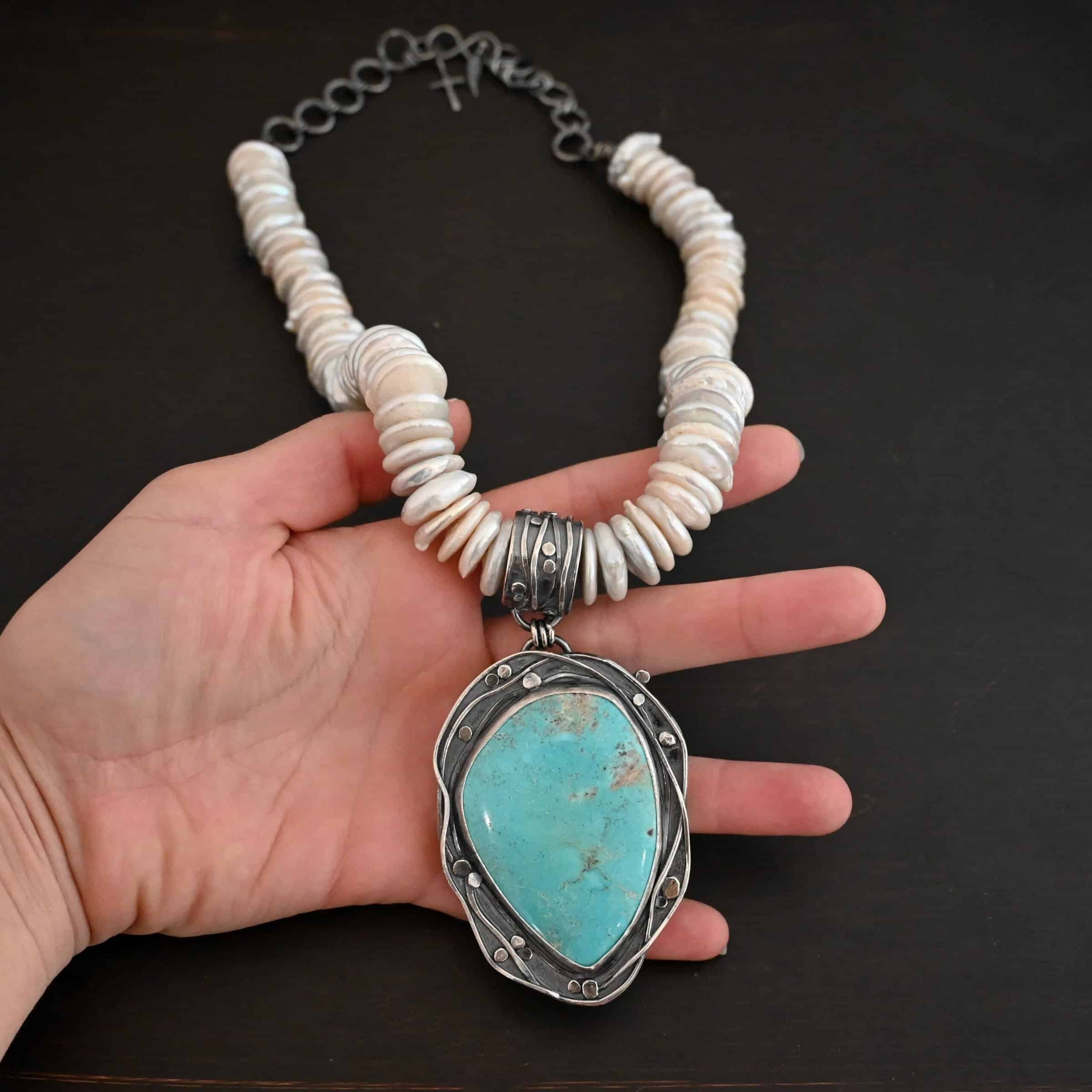 Calypso Necklace Photo of the "Calypso" Necklace. It is a sterling silver, turquoise, and pearl necklace. There is a large, oval shaped turquoise. It is set in sterling silver. The inside of the silver setting has hammered silver dots going all around it, and the outside of the silver setting has swirling silver lines going all around the turquoise. The collar of the necklace is made entirely out of strung pearl disks that are large and rough circle shapes. There is a large silver attachment on the top of the turquoise pendant to attach it to the collar. The attachment has a hole going through the center, and it is in the center of the necklace, with pearls on each side of it. Necklace is laying against the palm of a hand to demonstrate size.